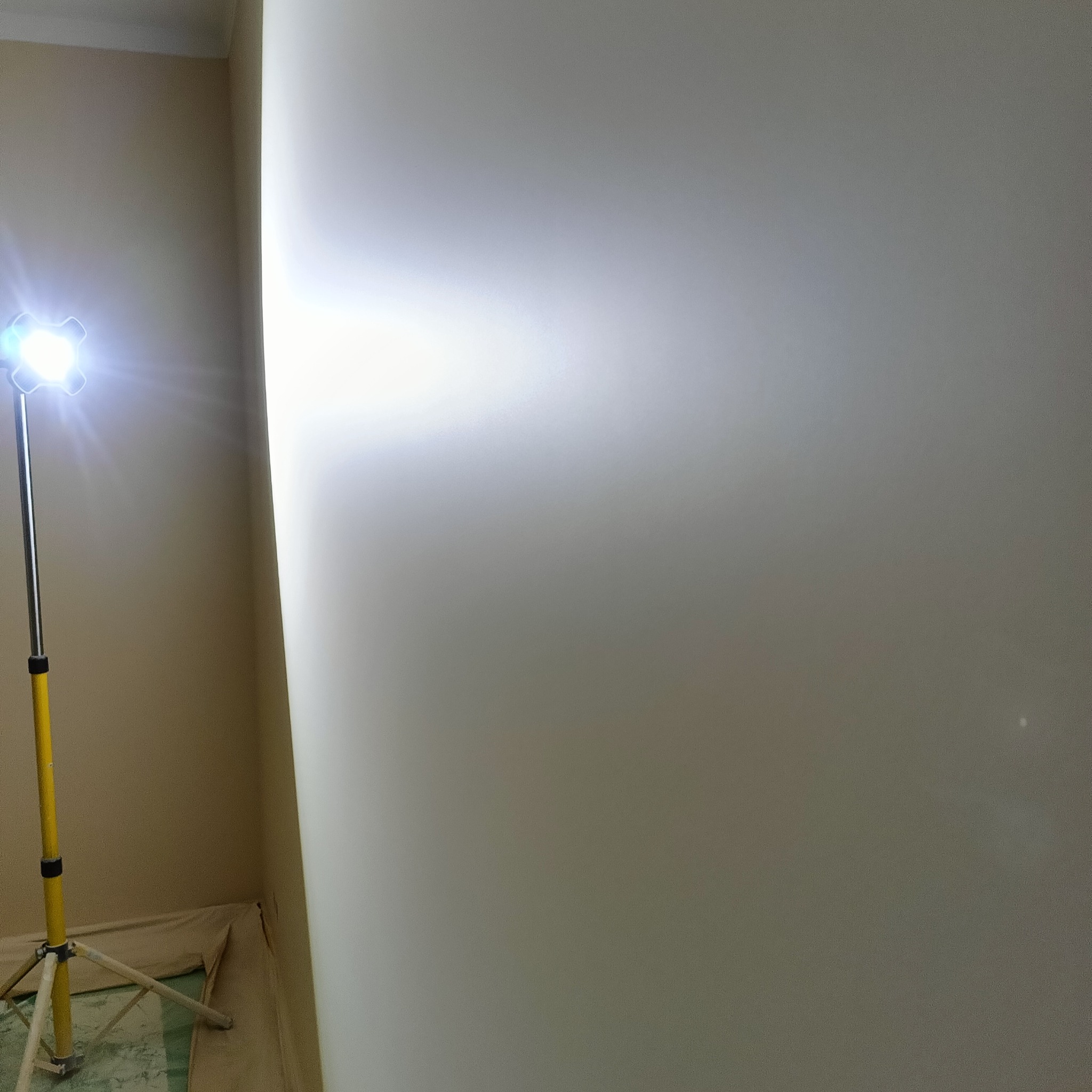 Ściana po gładziowaniu, oświetlona lampą budowlaną. Widoczny fragment zabezpieczonej podłogi. Świeżo wygładzona powierzchnia gotowa do malowania.