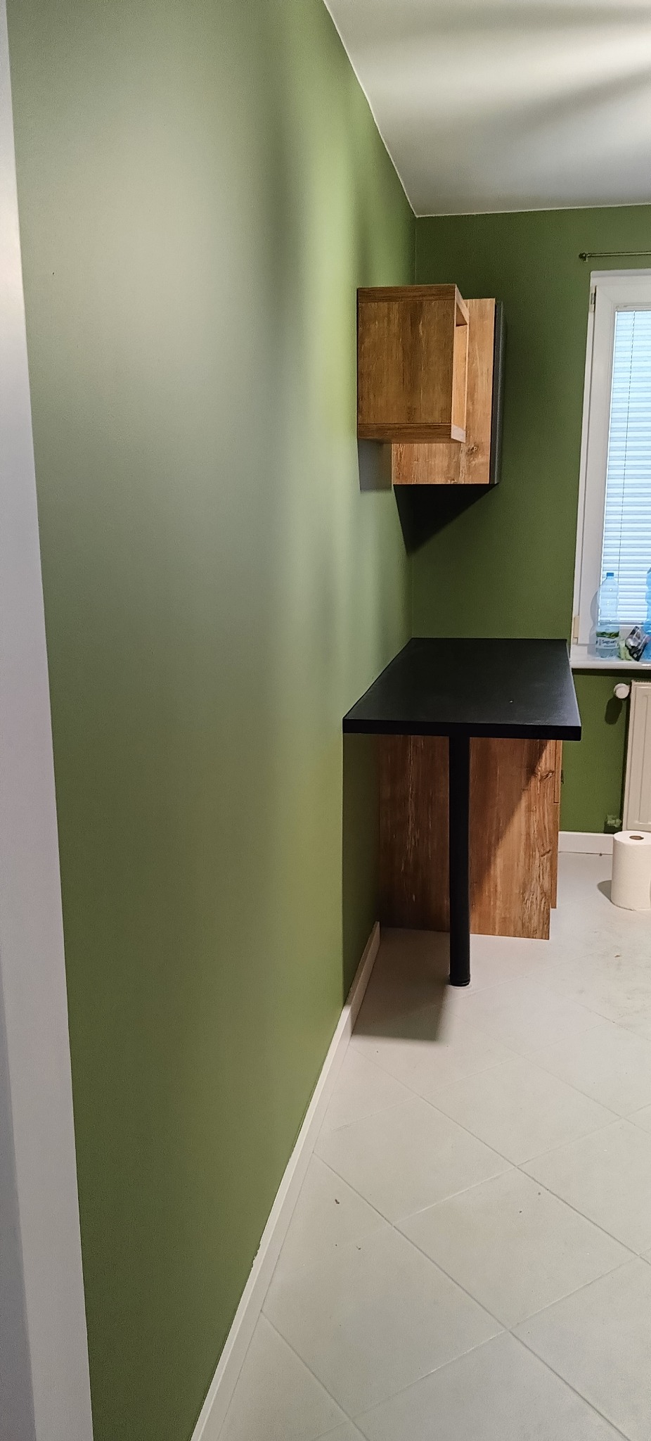 Nowoczesny pokój z biurkiem z czarnym blatem i wiszącą szafką z drewnianym frontem, na tle ściany w odcieniach zieleni. Minimalistyczny design, jasna podłoga.