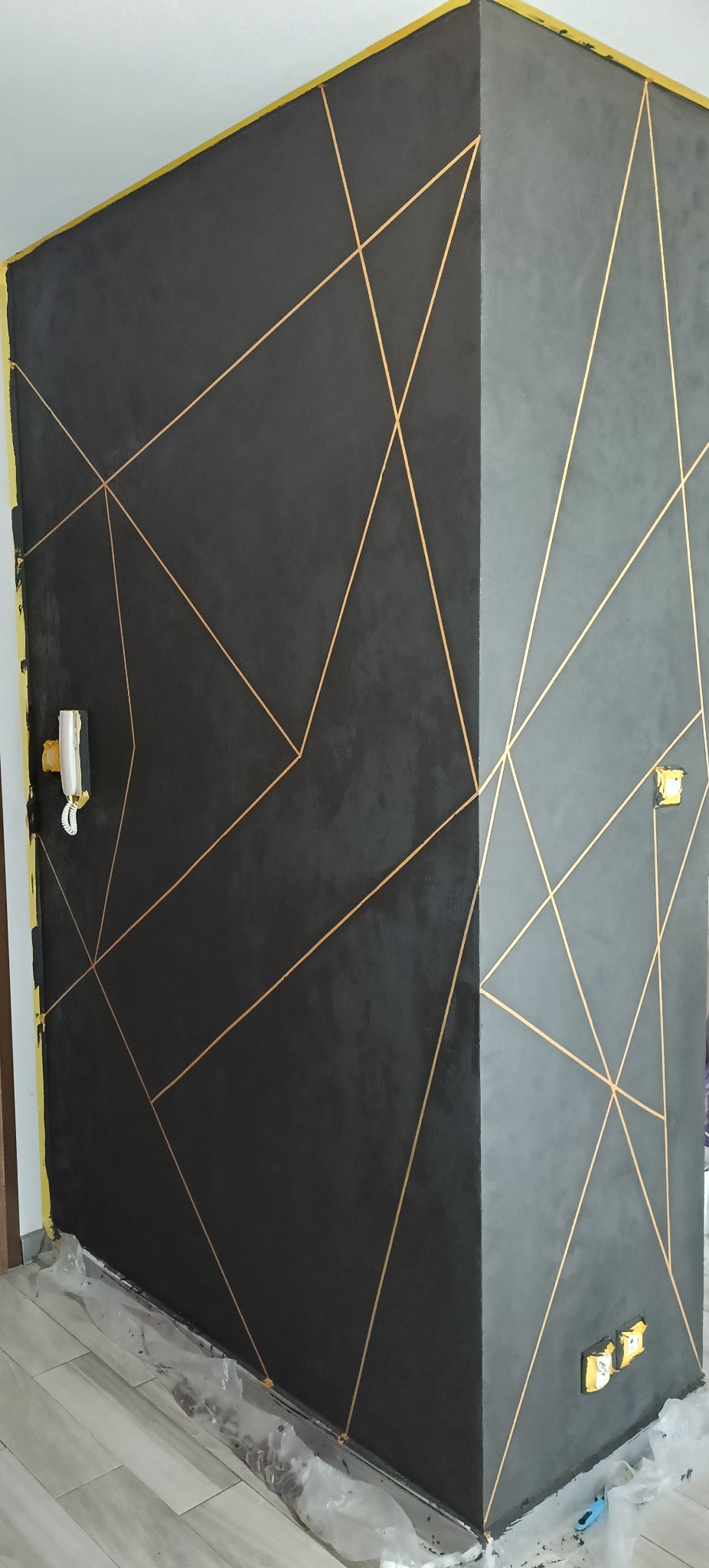 Ściana w odcieniach szarości z geometrycznym wzorem z cienkich, złotych linii. Widoczne zabezpieczenia podłogi i gniazdka elektryczne.