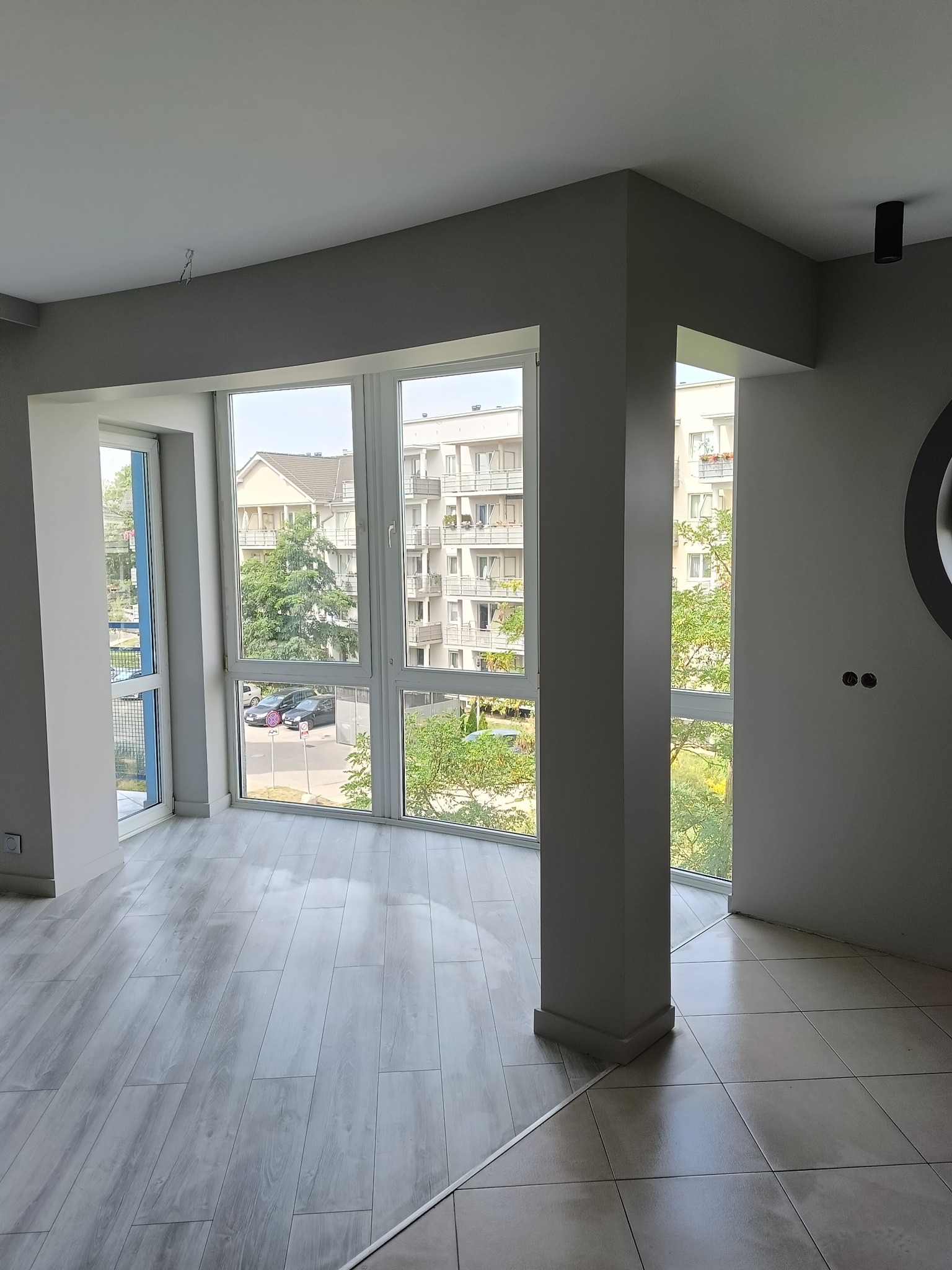 Jasne, pomalowane na szaro wnętrze z dużymi oknami i panelami podłogowymi. Widoczne gniazdka elektryczne i fragment lampy sufitowej. W tle budynek mieszkalny.