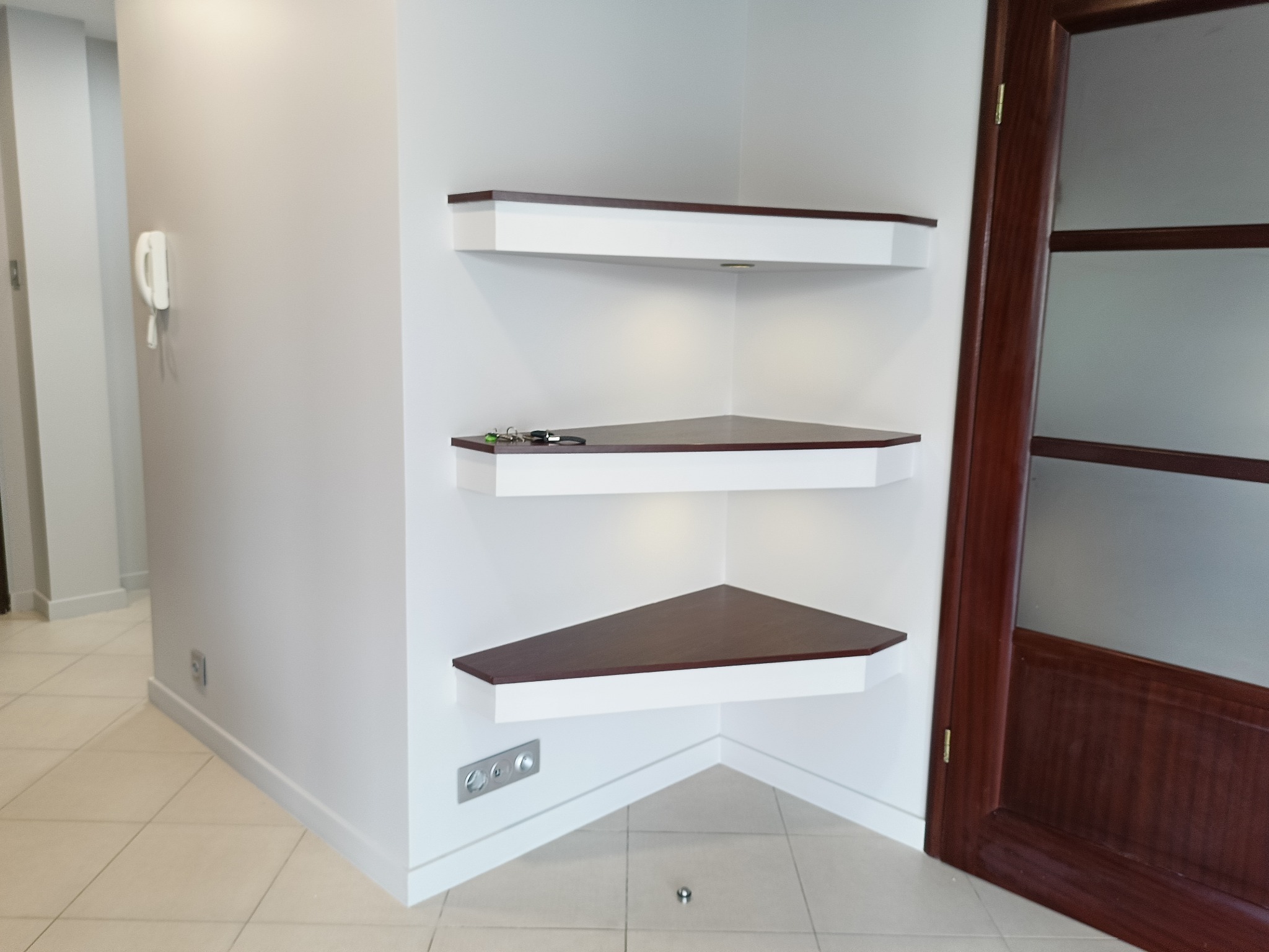 Kaskadowe, narożne półki z ciemnego drewna i białych wsporników, z oświetleniem punktowym, w jasnym wnętrzu z drewnianymi drzwiami i beżową podłogą.