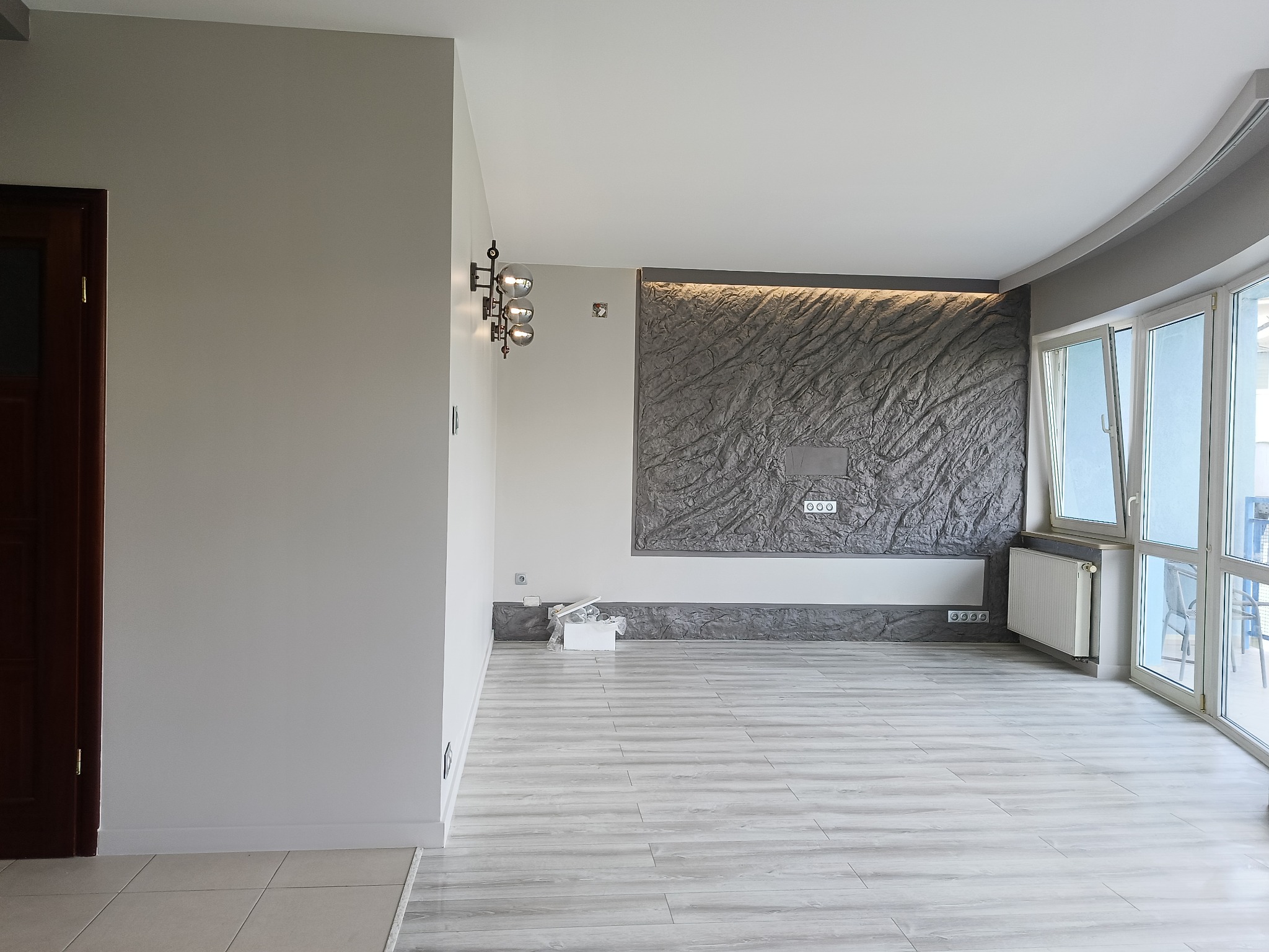 Jasne, przestronne wnętrze po remoncie z panelami podłogowymi, dekoracyjną ścianą z kamienia i nowoczesnym oświetleniem. Widok na balkon przez duże okna.
