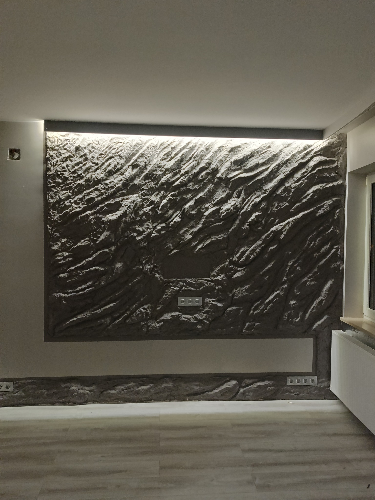 Ściana z efektem kamienia, podświetlona listwą LED, z gniazdkami elektrycznymi. Nowoczesne wykończenie wnętrza, szara kolorystyka, widok na fragment podłogi i okna.