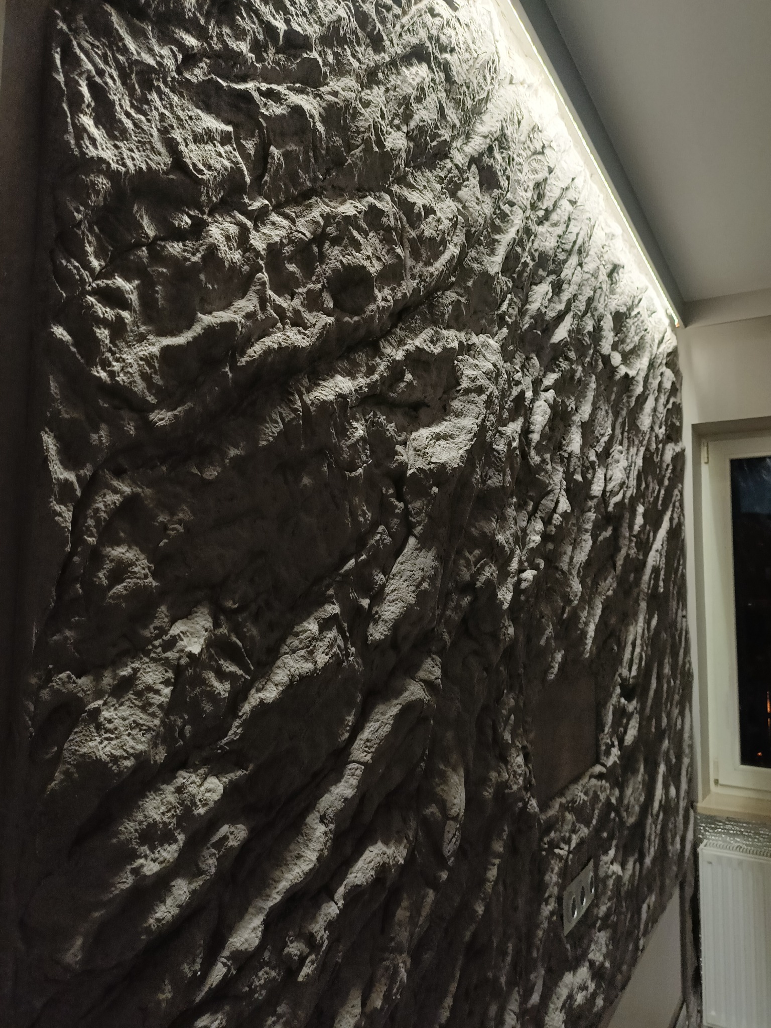 Ściana z dekoracyjną okładziną imitującą kamień, podświetlona listwą LED, z widocznymi gniazdkami elektrycznymi i fragmentem okna w tle.