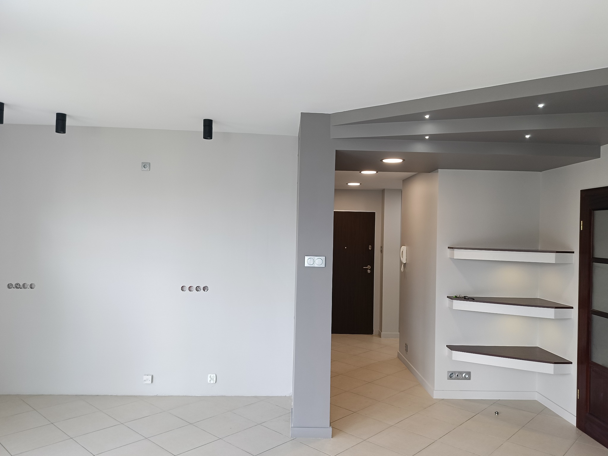 Jasne wnętrze po remoncie: szare ściany, płytki podłogowe, minimalistyczne półki narożne z oświetleniem, czarne reflektory sufitowe i gniazdka elektryczne.