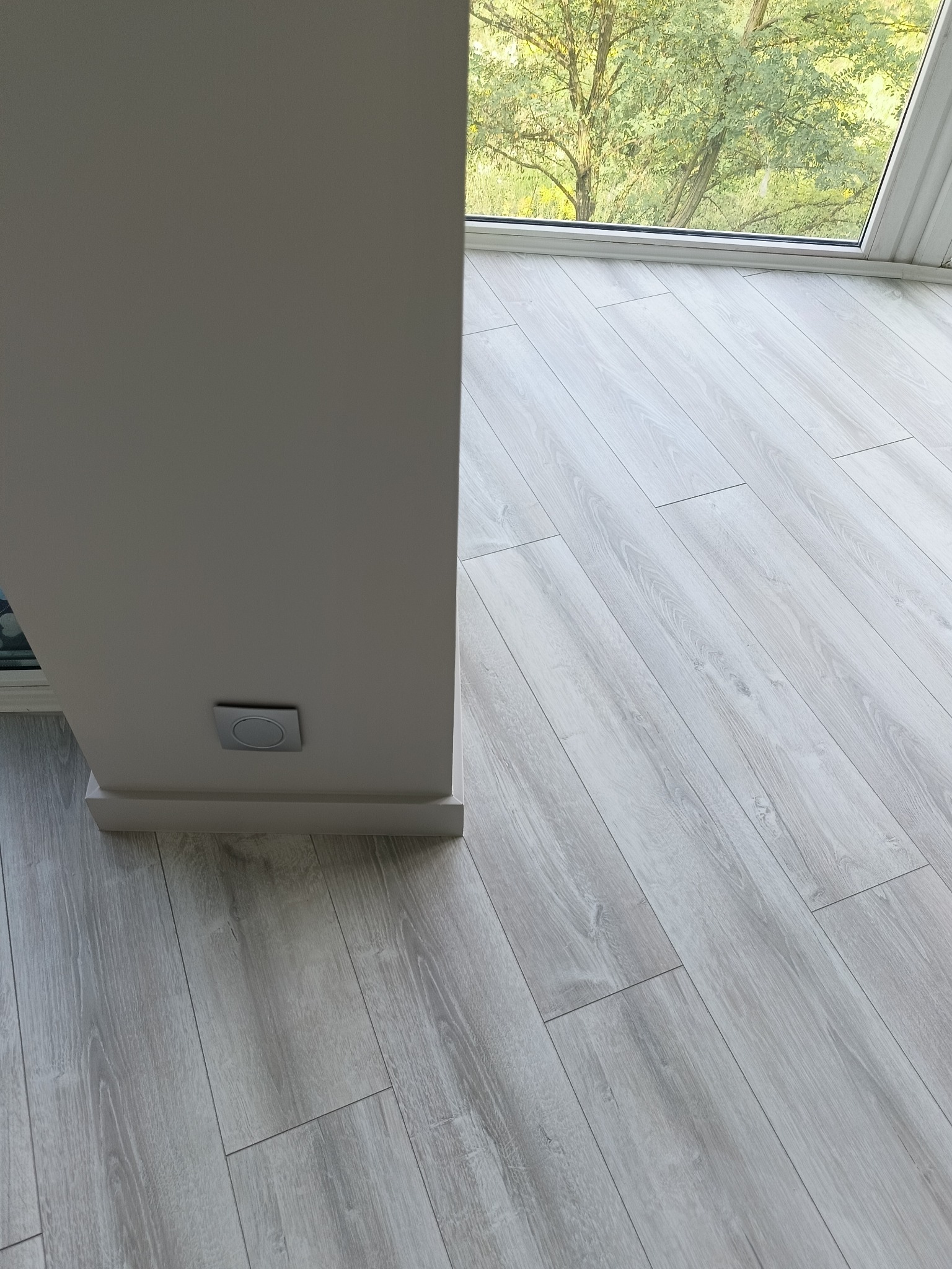 Jasne panele podłogowe ułożone wzdłuż okna z widokiem na drzewo. Widoczny fragment ściany z włącznikiem. Minimalistyczny design, neutralna kolorystyka.