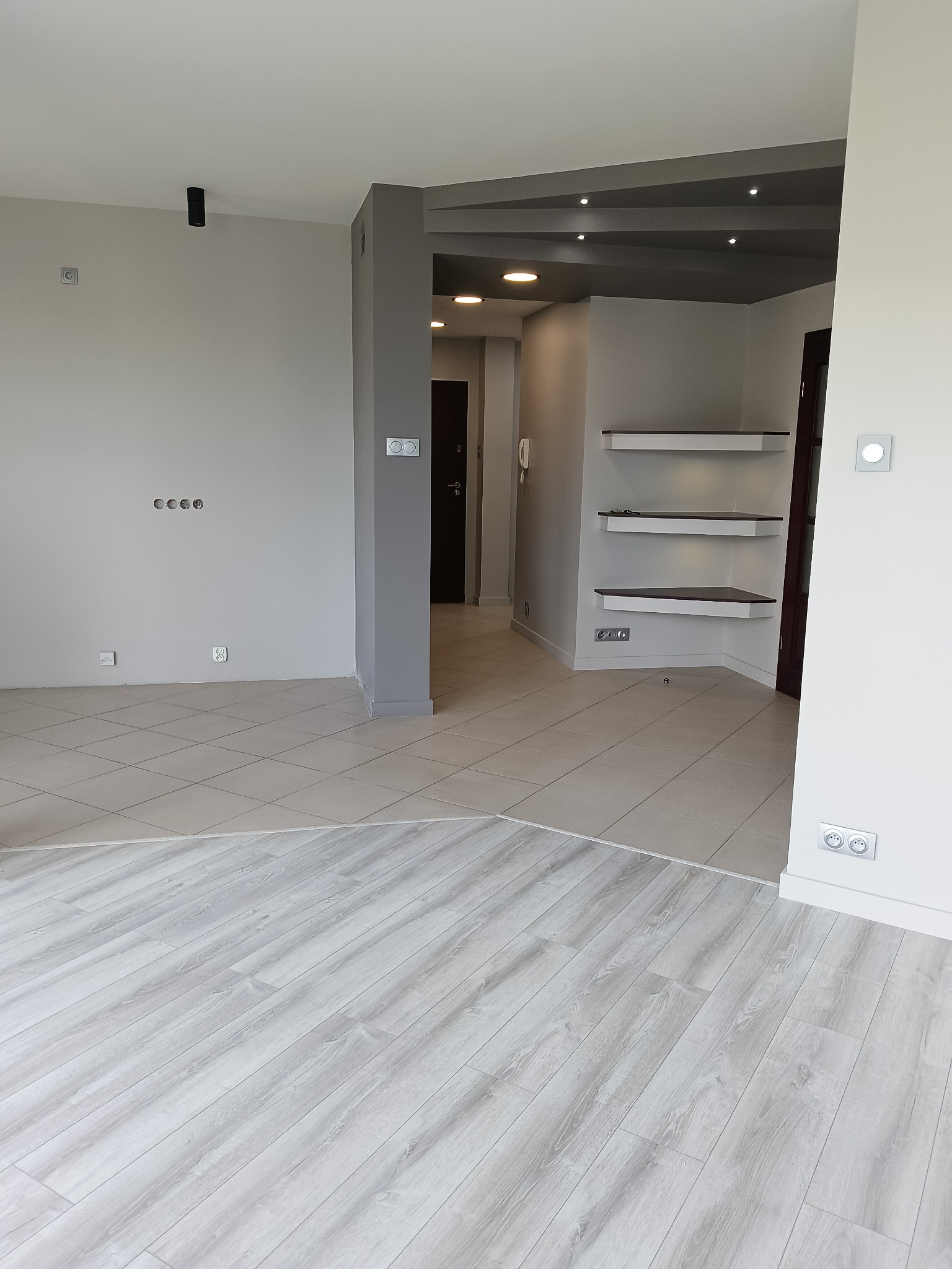 Jasne panele podłogowe przechodzą w beżowe płytki; widok na korytarz z szarym sufitem podwieszanym i półkami. Oświetlenie punktowe.