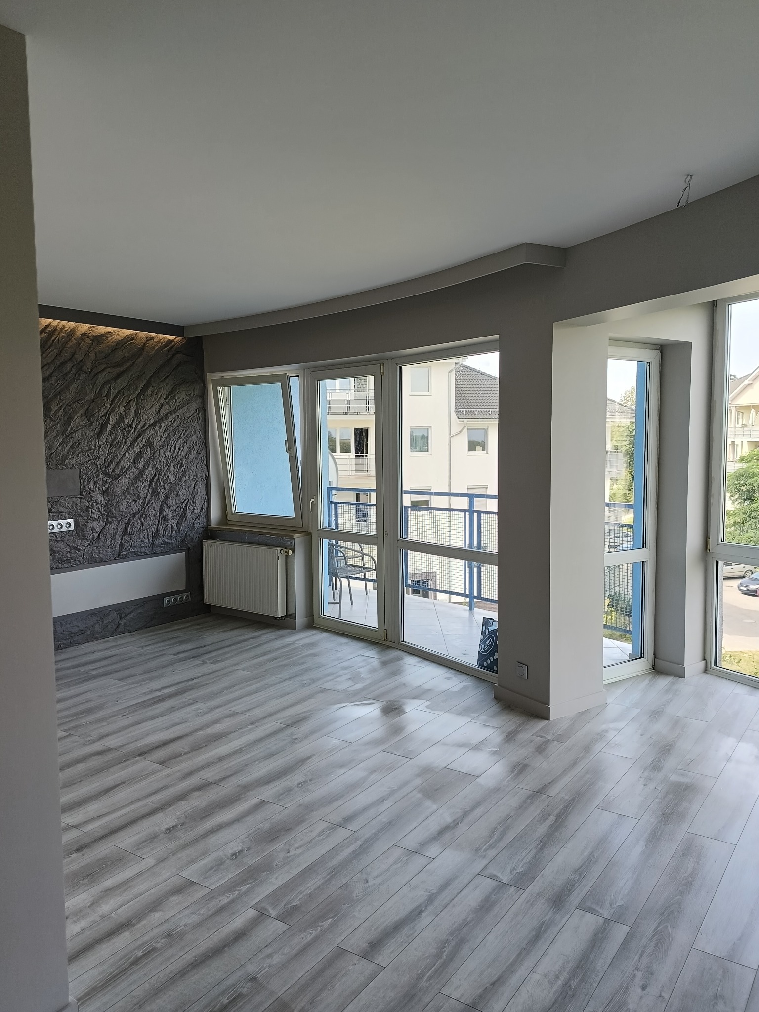 Nowoczesne wnętrze z szarą, teksturowaną ścianą, panelami podłogowymi i dużymi oknami balkonowymi z widokiem na zabudowę miejską. Minimalistyczny design i jasne, naturalne światło.