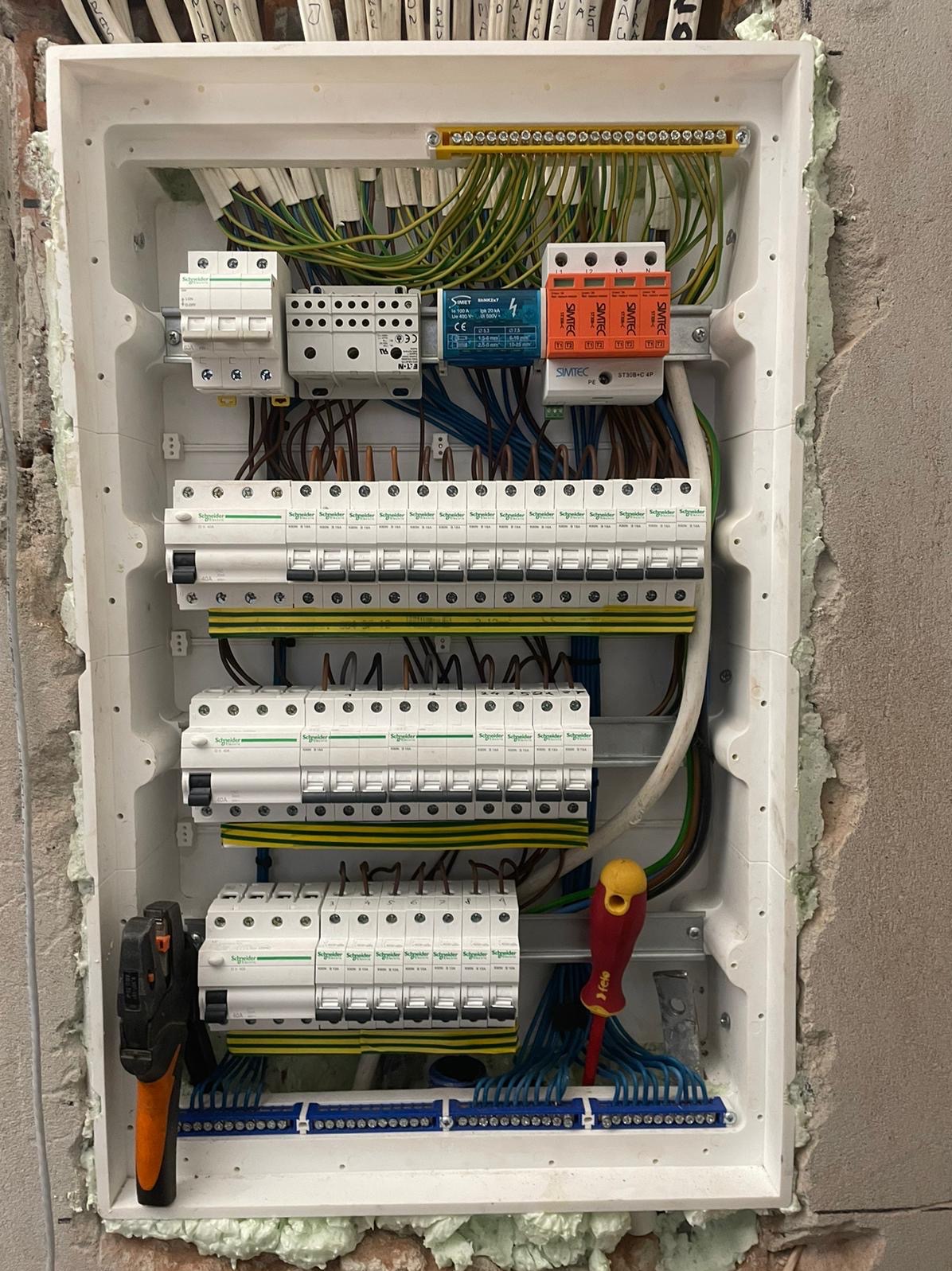 Rozdzielnica elektryczna w białej obudowie, z widocznymi bezpiecznikami, przewodami i narzędziami (szczypce, śrubokręt). Montaż w trakcie wykańczania, pianka montażowa wokół.