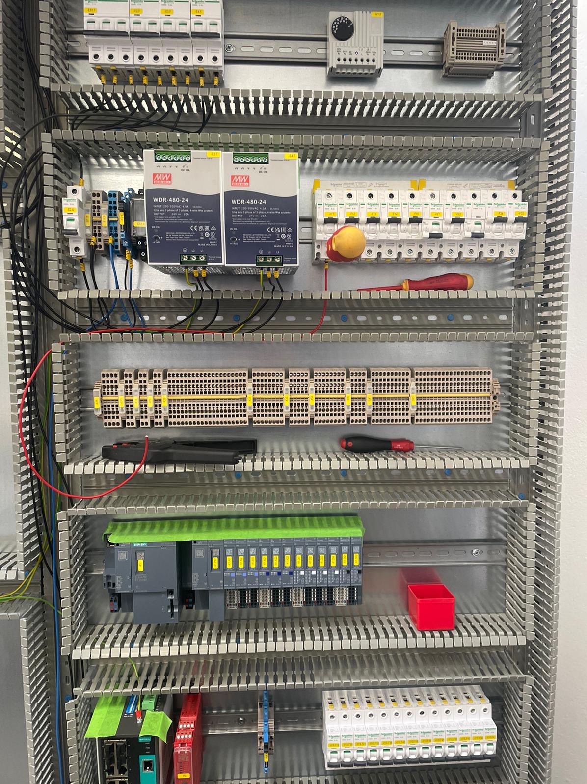Rozdzielnica elektryczna z modułami, zasilaczami MW WDR-480-24, bezpiecznikami i przewodami. Widoczne narzędzia: śrubokręty. Całość w metalowej obudowie.
