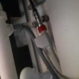 Szczegółowe ujęcie instalacji hydraulicznej pod zlewem, prezentujące podłączenie z zaworem z czerwoną nakrętką, węże odpływowe i dopływowe. Ujęcie od dołu.