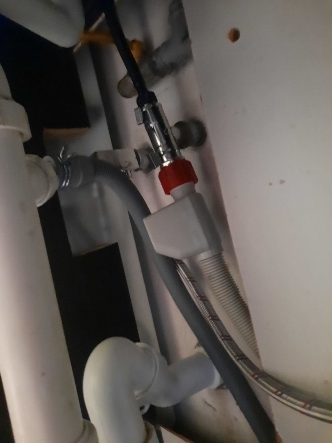 Szczegółowe ujęcie instalacji hydraulicznej pod zlewem, prezentujące podłączenie z zaworem z czerwoną nakrętką, węże odpływowe i dopływowe. Ujęcie od dołu.