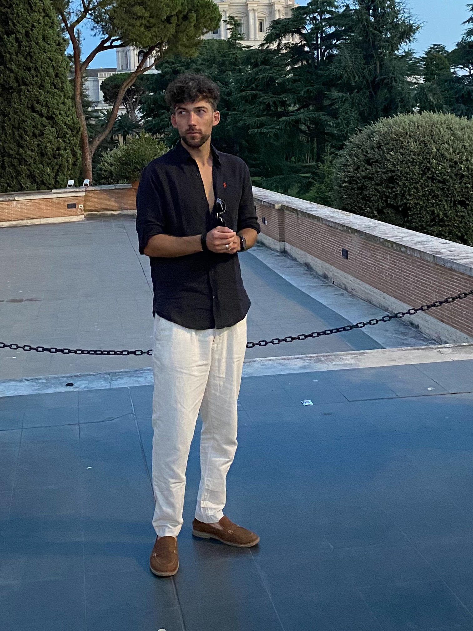 Mężczyzna w czarnej koszuli i lnianych spodniach stoi na tle muru z łańcuchem i zieleni miejskiej. W tle budynek z kopułą.