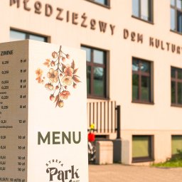 Strefa Park sp. z o.o. - Eleganckie menu 'Strefa Park' z cennikiem na tle budynku Młodzieżowego Domu Kultury. Kwiatowy motyw dekoracyjny, ciepłe oświetlenie.