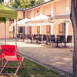 Strefa Park sp. z o.o. - Letni ogródek restauracji z parasolami Coca-Cola, drewnianymi leżakami i stolikami z krzesłami. Ujęcie z perspektywy trawnika, w tle budynek restauracji.