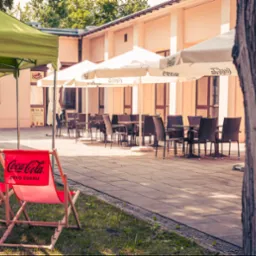 Letni ogr&oacute;dek restauracji z parasolami Coca-Cola, drewnianymi leżakami i stolikami z krzesłami. Ujęcie z perspektywy trawnika, w tle budynek restauracji.