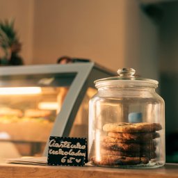 Strefa Park sp. z o.o. - Słoik z czekoladowymi ciastkami obok tabliczki z ceną w cukierni. W tle witryna chłodnicza z wypiekami, ciepłe oświetlenie.