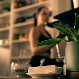 Strefa Park sp. z o.o. - Kawiarnia: roślina w doniczce, serwetki na talerzyku, kobieta w tle przy ekspresie. Klimatyczne oświetlenie, ciepła atmosfera. Detal wnętrza kawiarni.