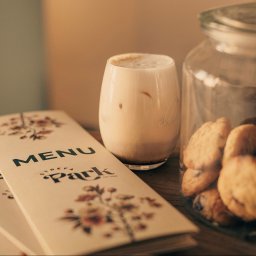 Strefa Park sp. z o.o. - Stylowe menu z kwiatowym motywem obok szklanki latte i słoika ciasteczek na drewnianym stole, ciepłe oświetlenie, klimatyczna kawiarnia.