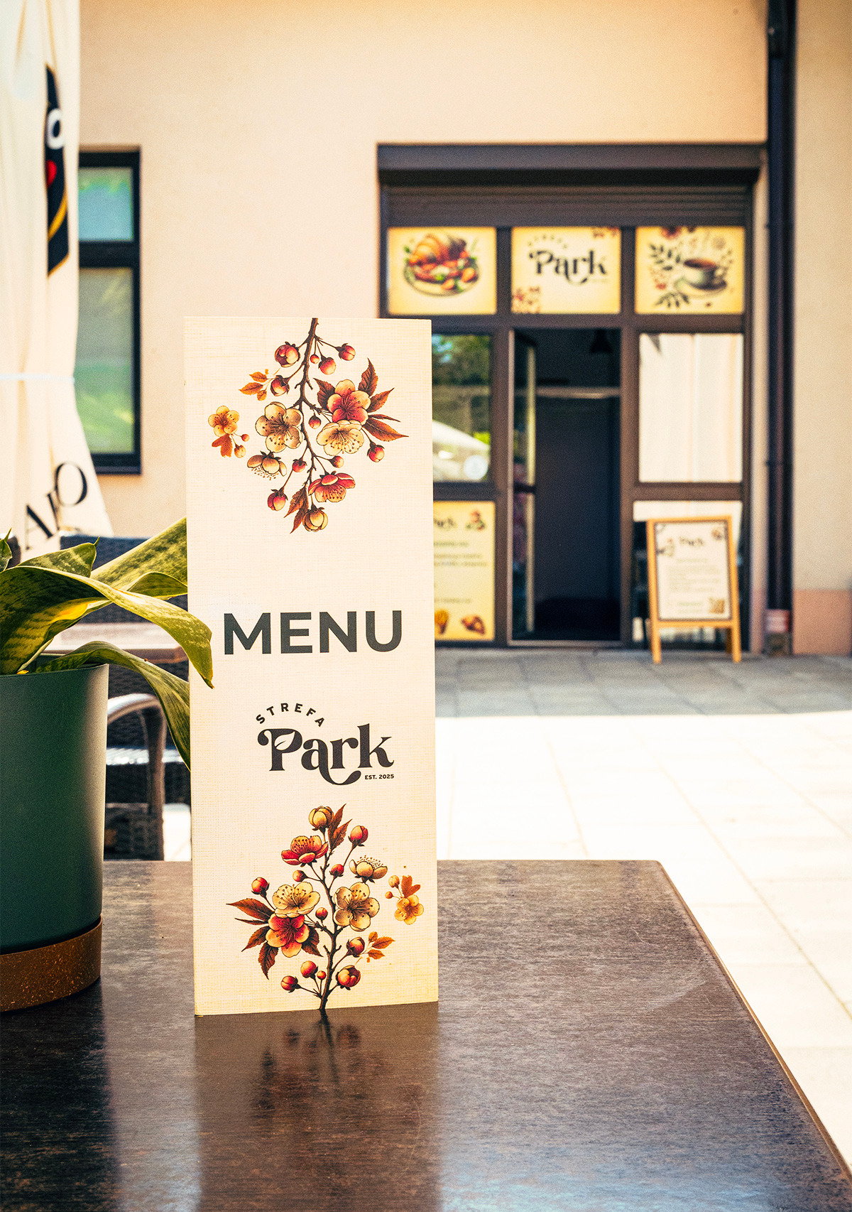Menu restauracji 'Strefa Park' z motywem kwiatowym na stoliku, z widokiem na wejście do lokalu i tablicę z ofertą na zewnątrz. Słoneczny dzień.