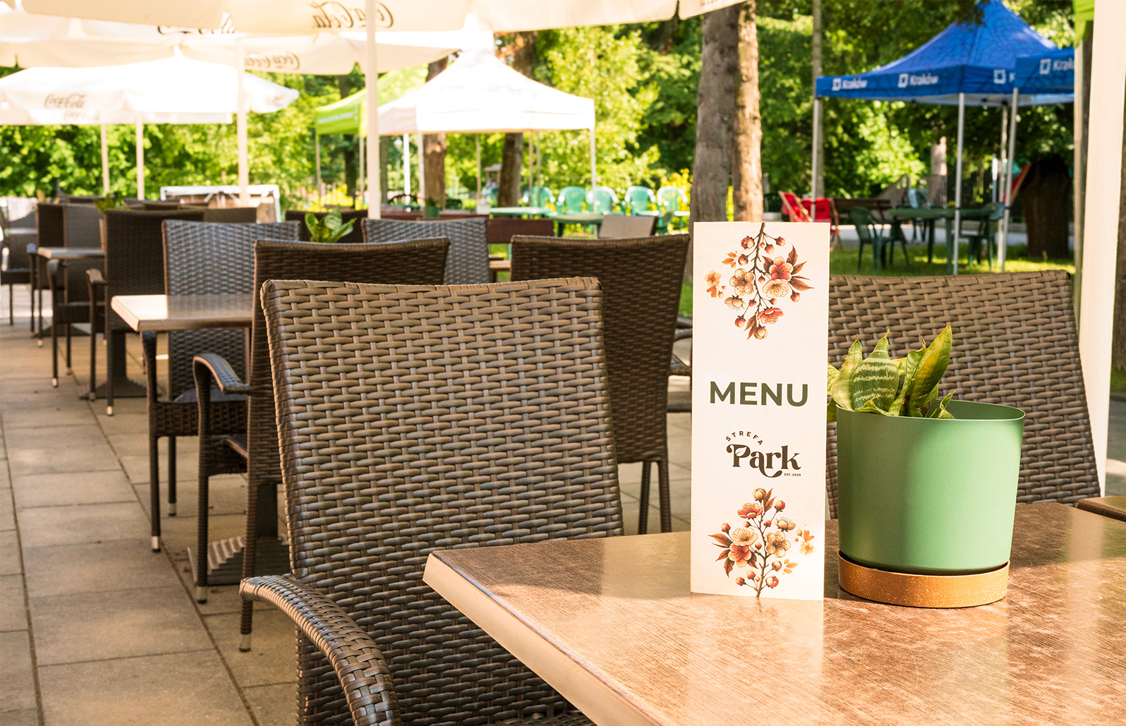 Letni ogródek restauracji z rattanowymi krzesłami i stolikami. Na stole menu z kwiatowym motywem i zielona roślina w doniczce. W tle parasole i zieleń.