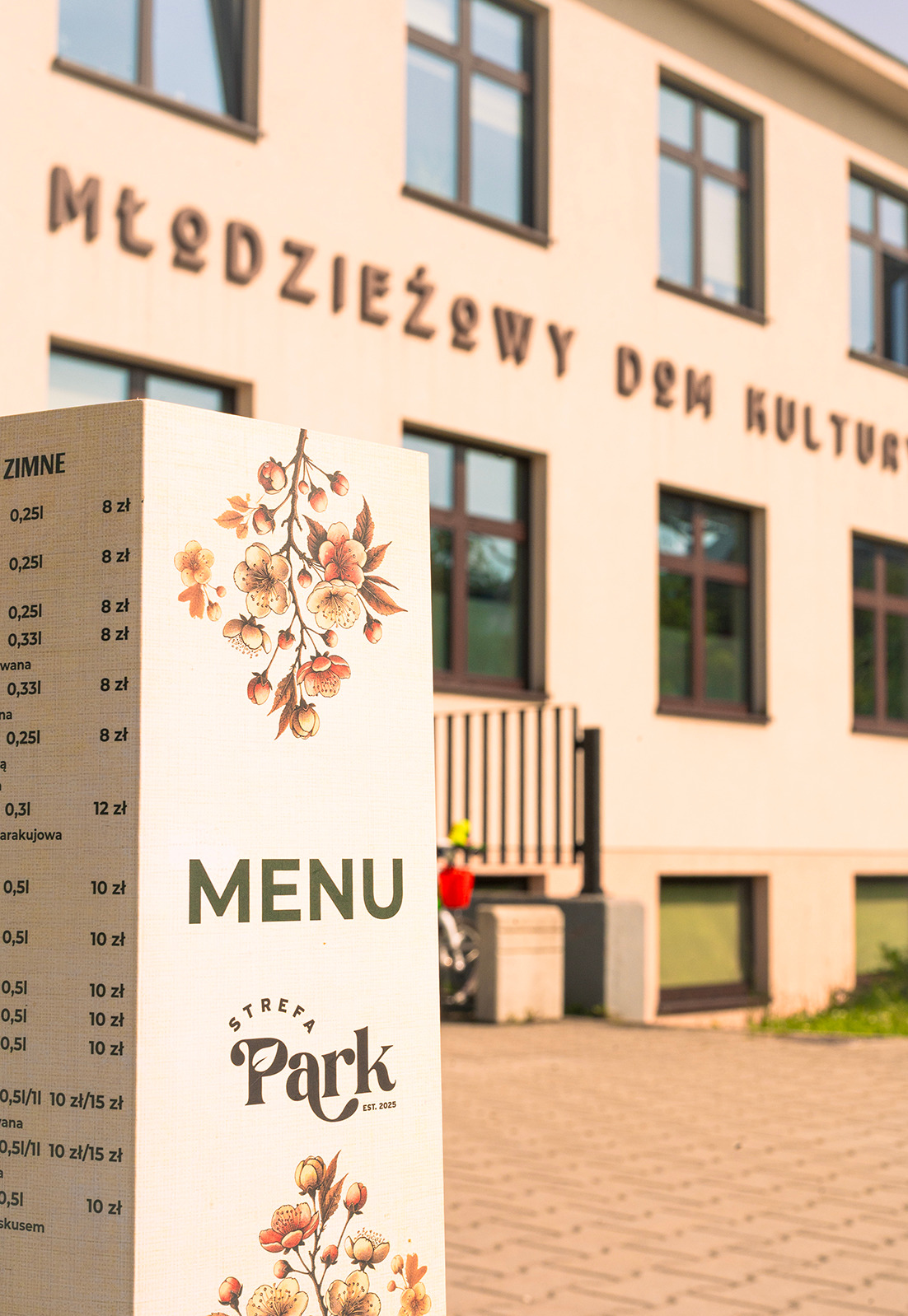 Eleganckie menu 'Strefa Park' z cennikiem na tle budynku Młodzieżowego Domu Kultury. Kwiatowy motyw dekoracyjny, ciepłe oświetlenie.