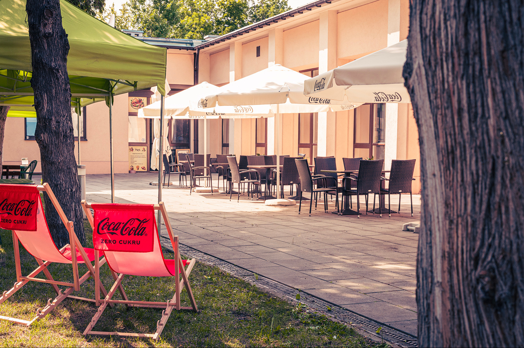 Letni ogródek restauracji z parasolami Coca-Cola, drewnianymi leżakami i stolikami z krzesłami. Ujęcie z perspektywy trawnika, w tle budynek restauracji.