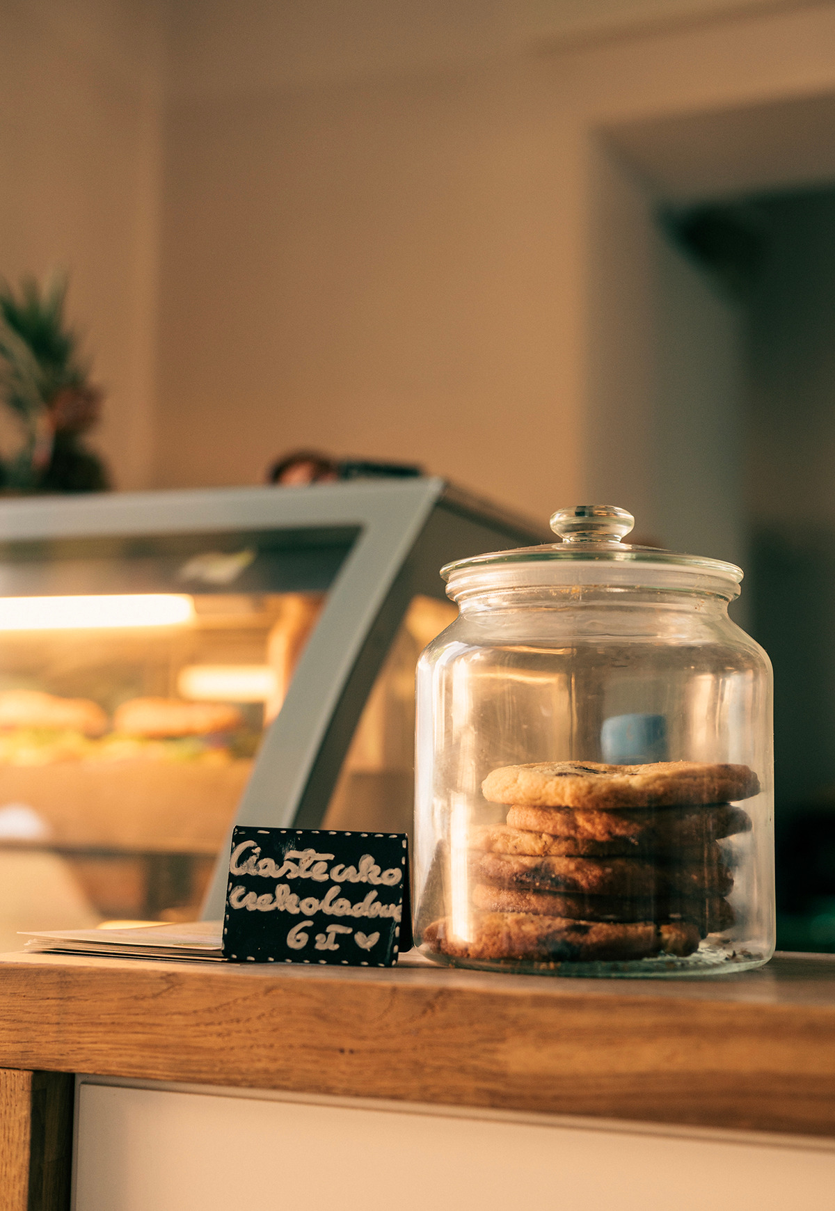 Słoik z czekoladowymi ciastkami obok tabliczki z ceną w cukierni. W tle witryna chłodnicza z wypiekami, ciepłe oświetlenie.