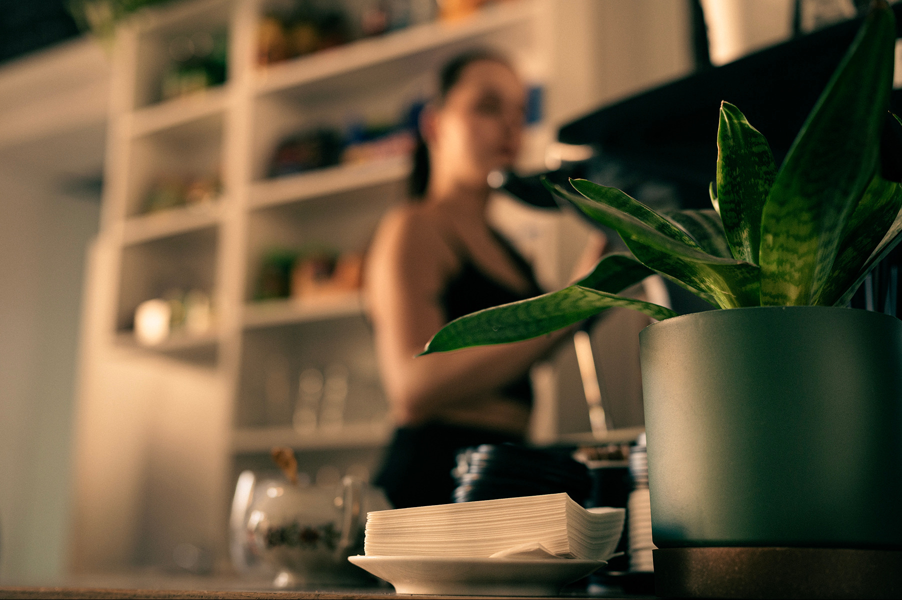 Kawiarnia: roślina w doniczce, serwetki na talerzyku, kobieta w tle przy ekspresie. Klimatyczne oświetlenie, ciepła atmosfera. Detal wnętrza kawiarni.