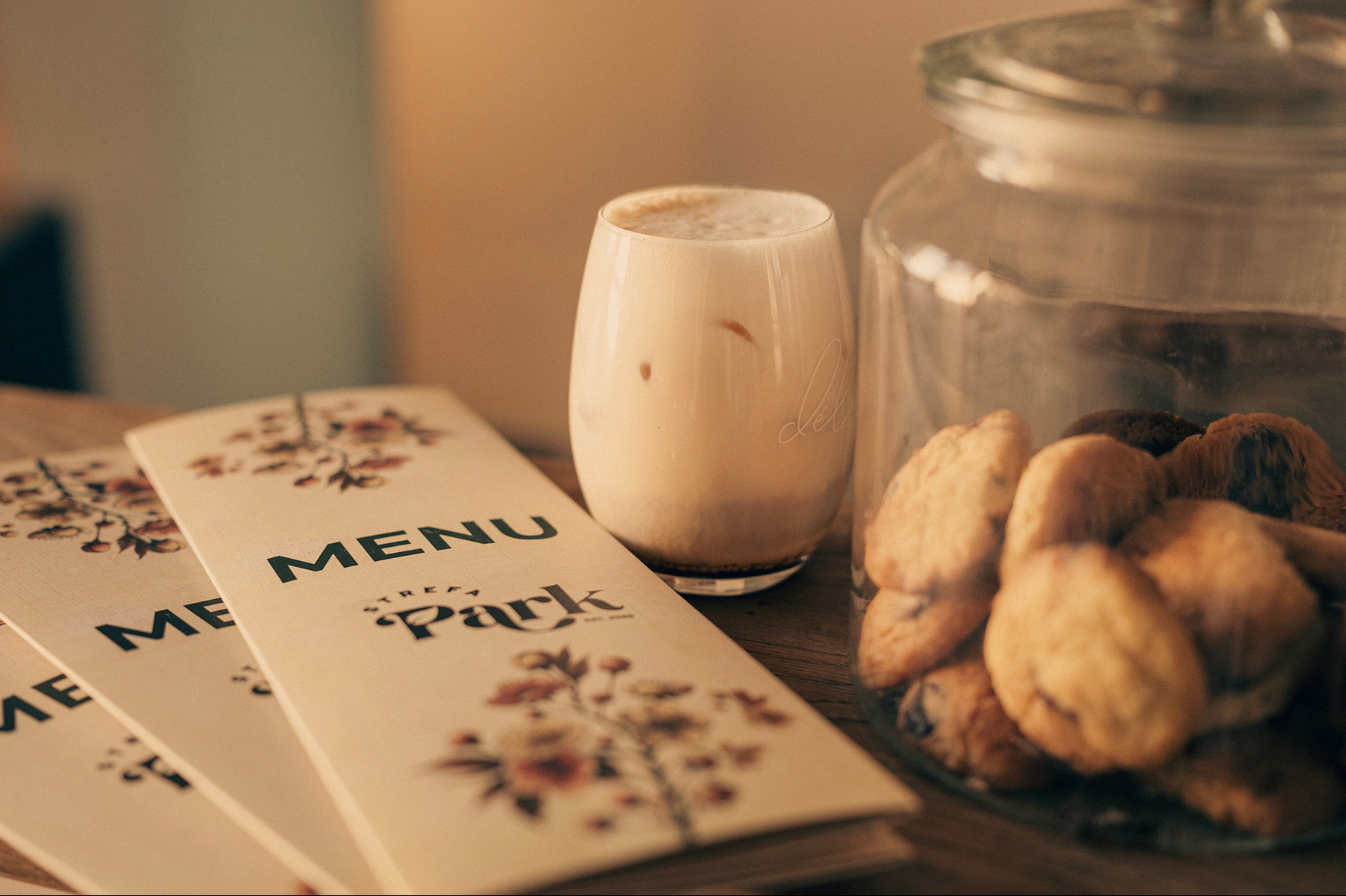 Stylowe menu z kwiatowym motywem obok szklanki latte i słoika ciasteczek na drewnianym stole, ciepłe oświetlenie, klimatyczna kawiarnia.