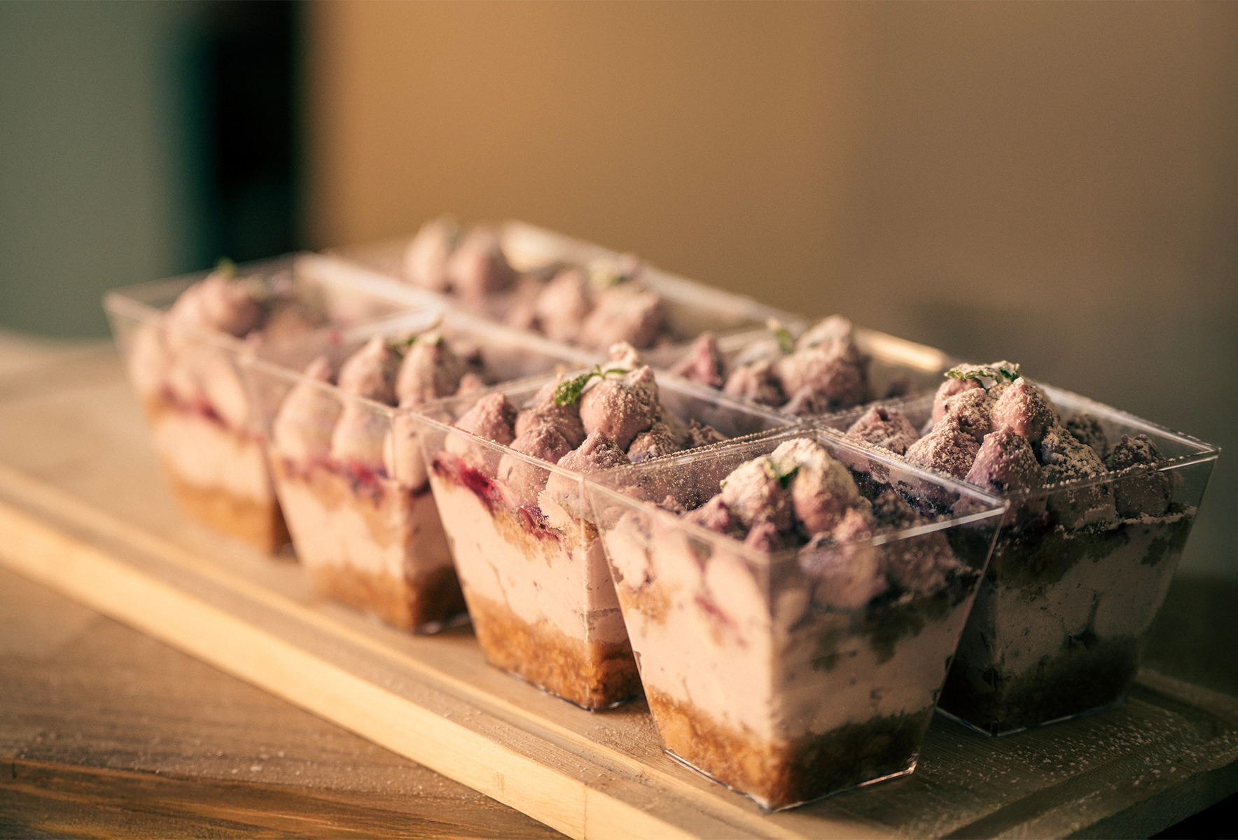 Deserki w przezroczystych pojemnikach, ułożone na drewnianej desce. Kremowe desery z owocami i posypką, gotowe do podania, w ciepłej tonacji.