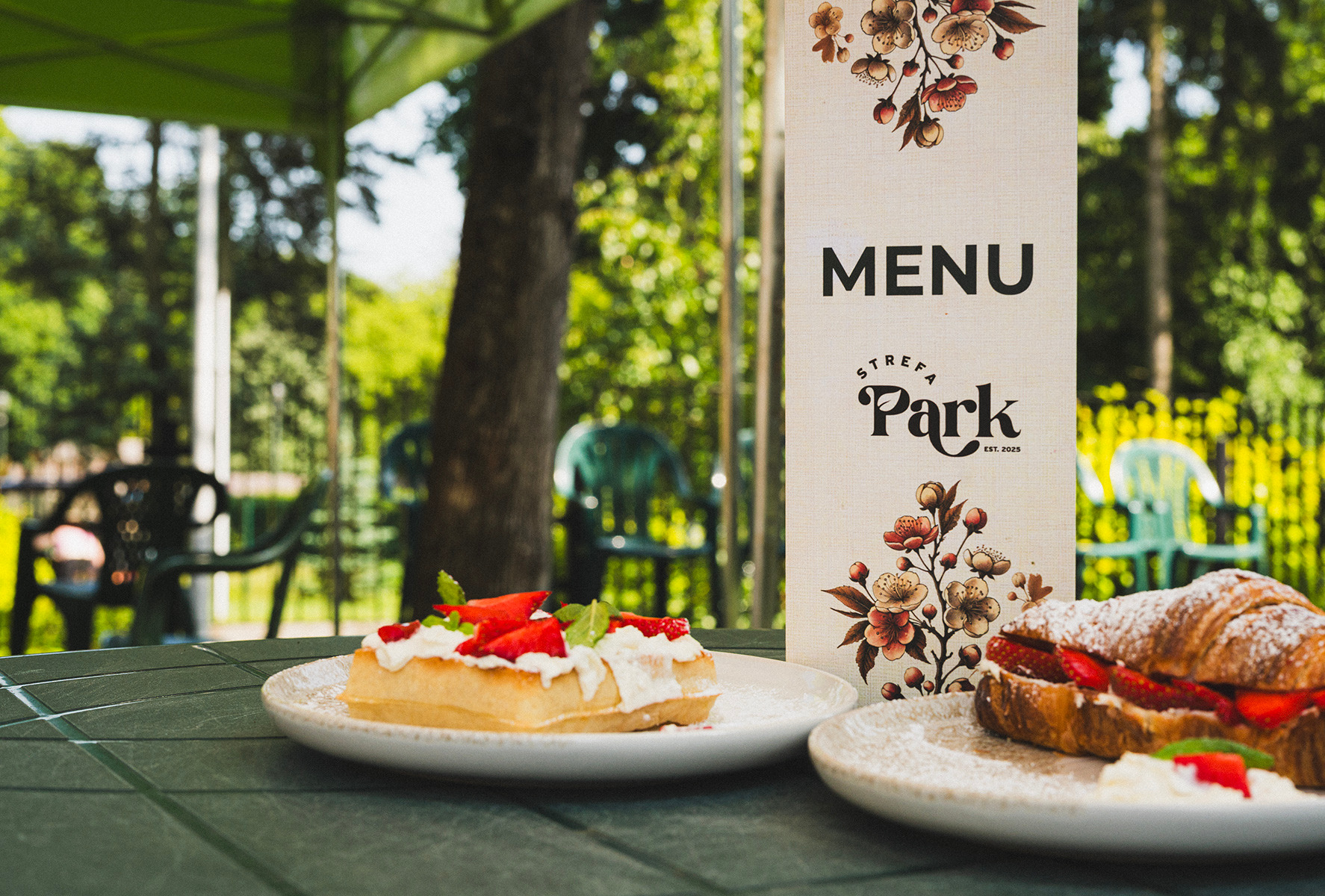 Letnia oferta kawiarni 'Strefa Park': gofr z bitą śmietaną i truskawkami obok menu z motywem kwiatowym, croissant z truskawkami w tle, zielony ogródek.