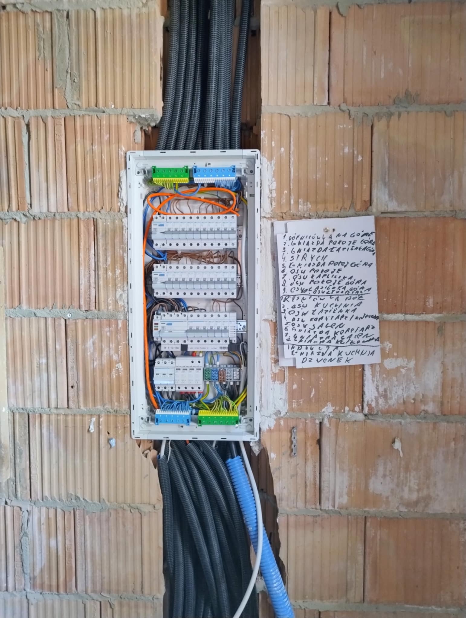 Rozdzielnia elektryczna w trakcie montażu w surowej ścianie z cegły, widoczne przewody i schemat instalacji na kartce. Instalacja w trakcie prac.