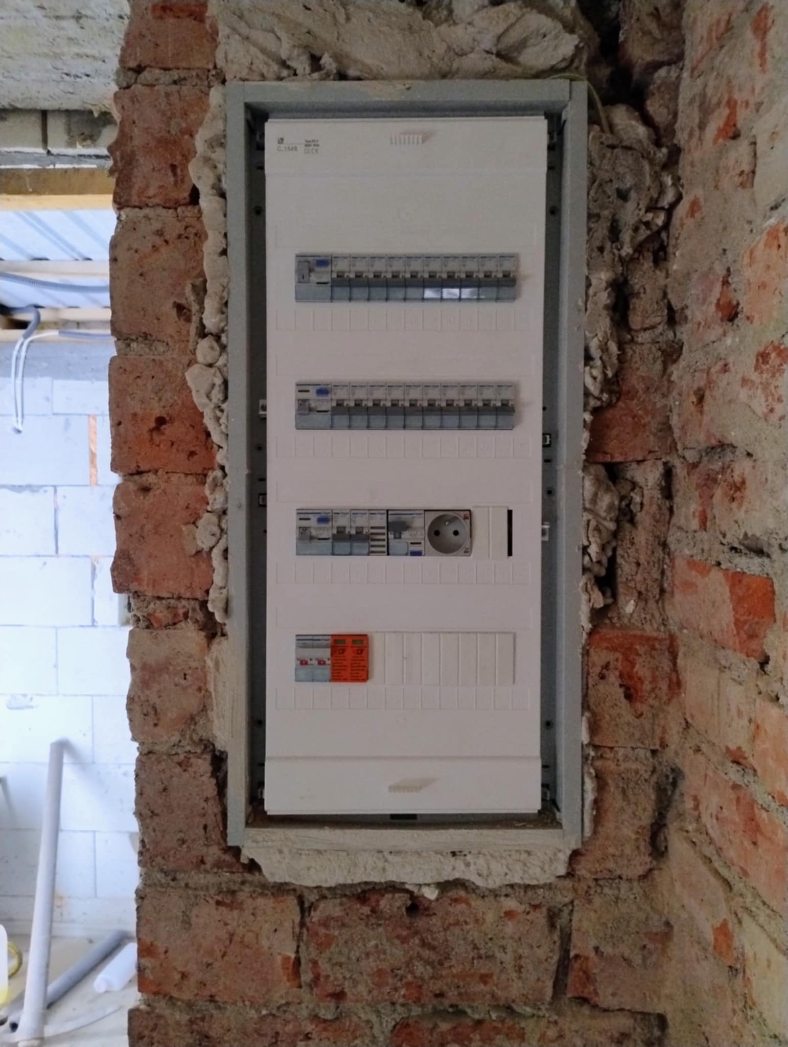 Rozdzielnica elektryczna zamontowana w surowej ścianie z czerwonej cegły, widoczne kable i zabezpieczenia, etap prac wykończeniowych.