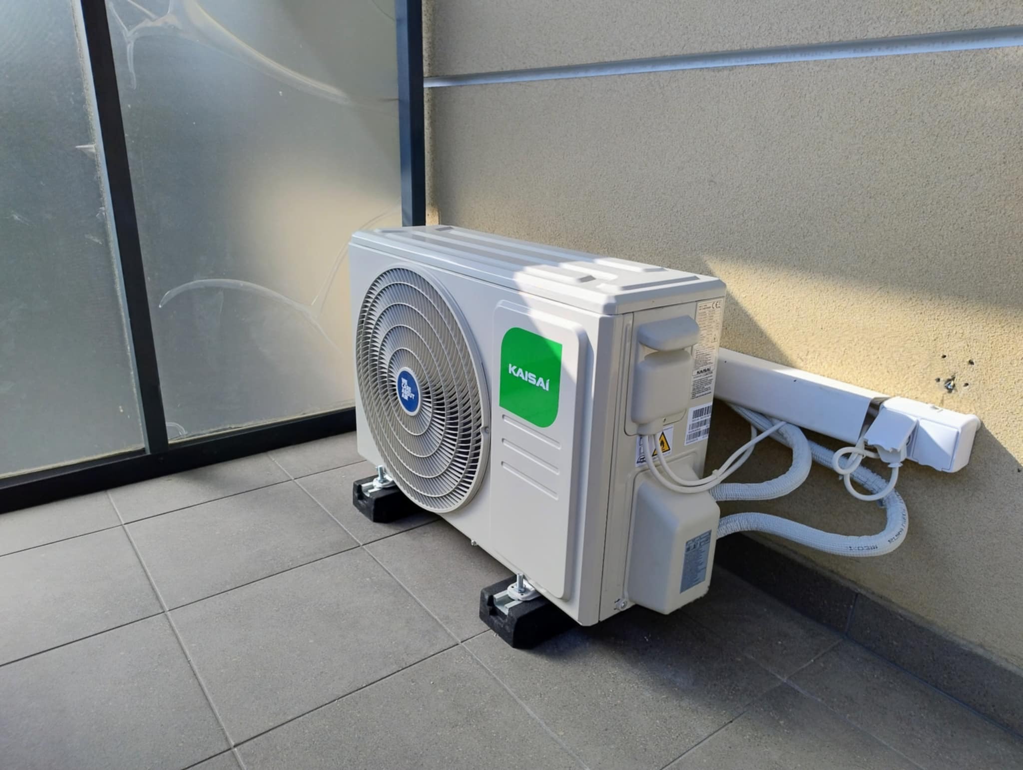 Biała jednostka zewnętrzna klimatyzacji Kaisai zamontowana na balkonie z szarą podłogą i beżową ścianą. Widoczne przewody i uchwyty montażowe.