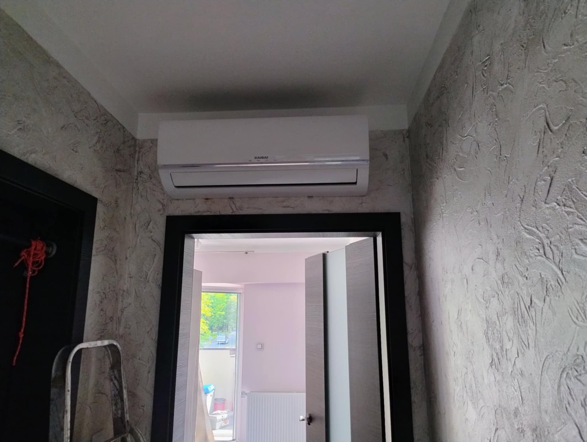 Biały klimatyzator Kaisai na ścianie z dekoracyjnym tynkiem, widok z korytarza na otwarte drzwi balkonowe. Widoczna drabina malarska.