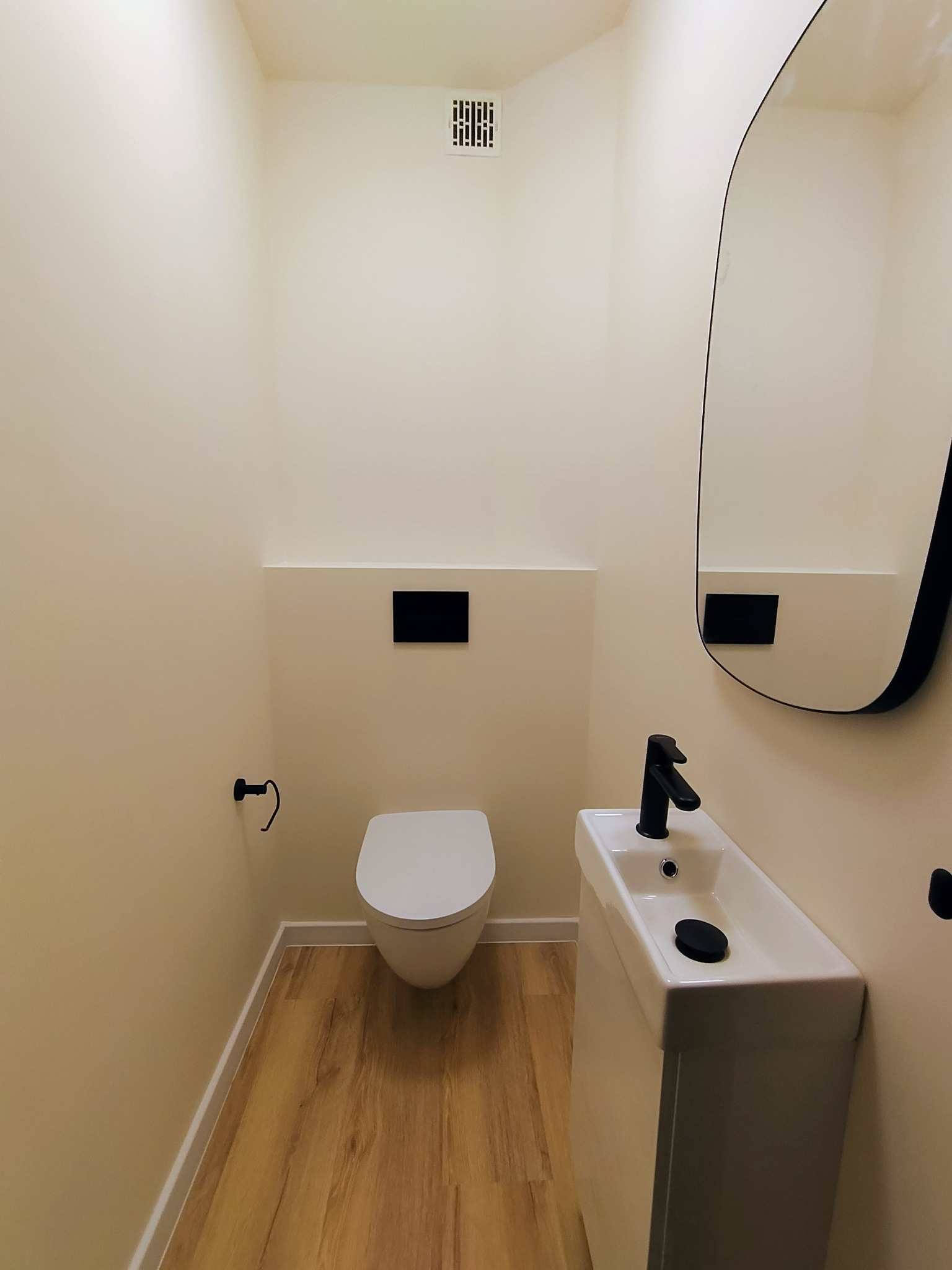 Nowoczesna toaleta w beżowej tonacji z drewnopodobną podłogą, wiszącą muszlą, umywalką na postumencie i owalnym lustrem z czarną ramą.