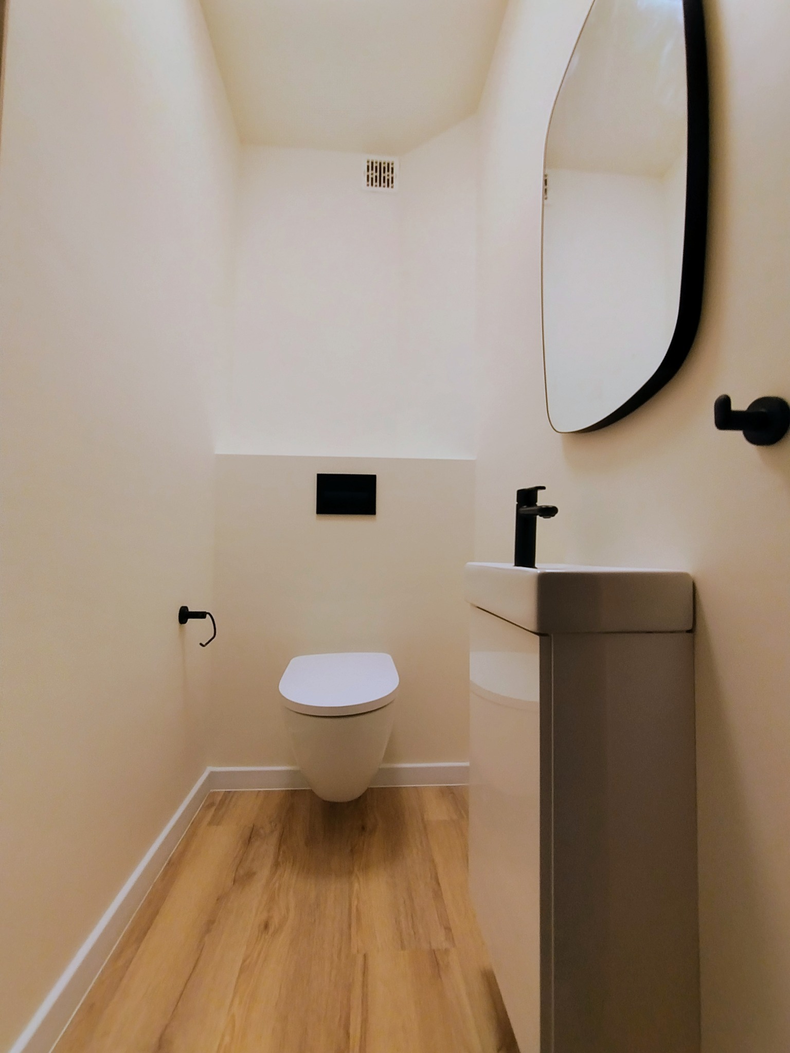 Nowoczesna toaleta w beżach z drewnianą podłogą, czarną armaturą i asymetrycznym lustrem. Minimalistyczny design, eleganckie wykończenie.