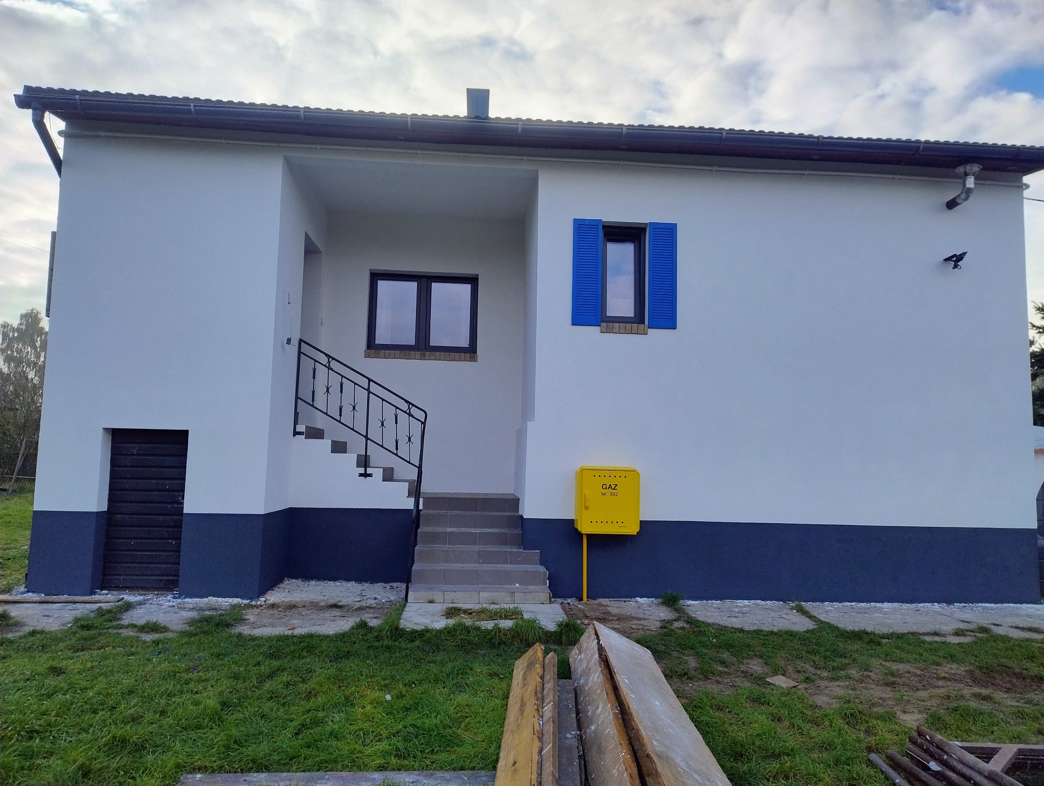 Biały dom z niebieskimi okiennicami i ciemnoszarą podmurówką. Żółta skrzynka gazowa przy wejściu. Schody z czarną balustradą prowadzą do drzwi.