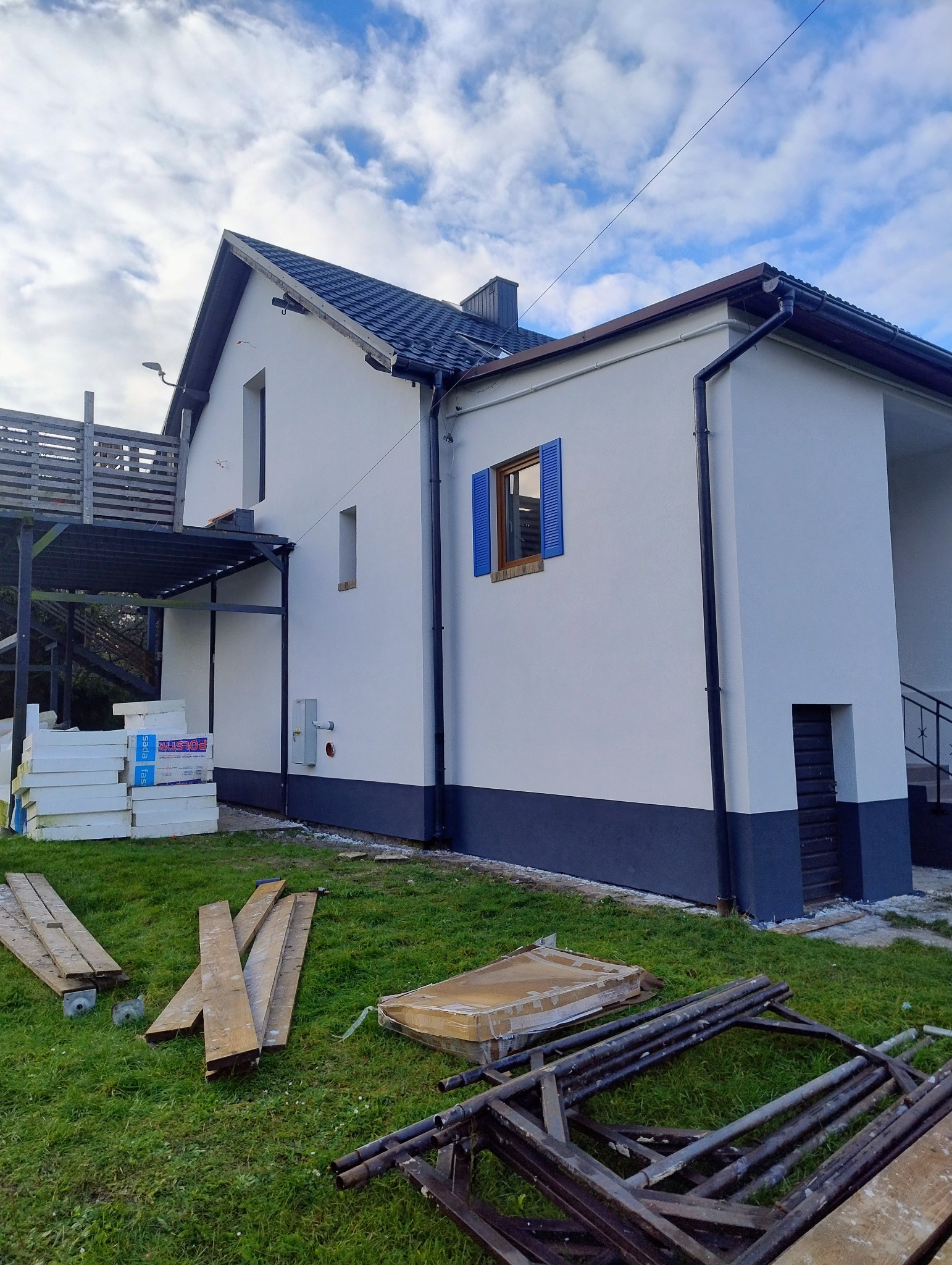Ocieplony dom jednorodzinny z niebieskimi okiennicami, widok z boku. Na trawie leżą materiały izolacyjne i rusztowanie. Błękitne niebo z chmurami w tle.