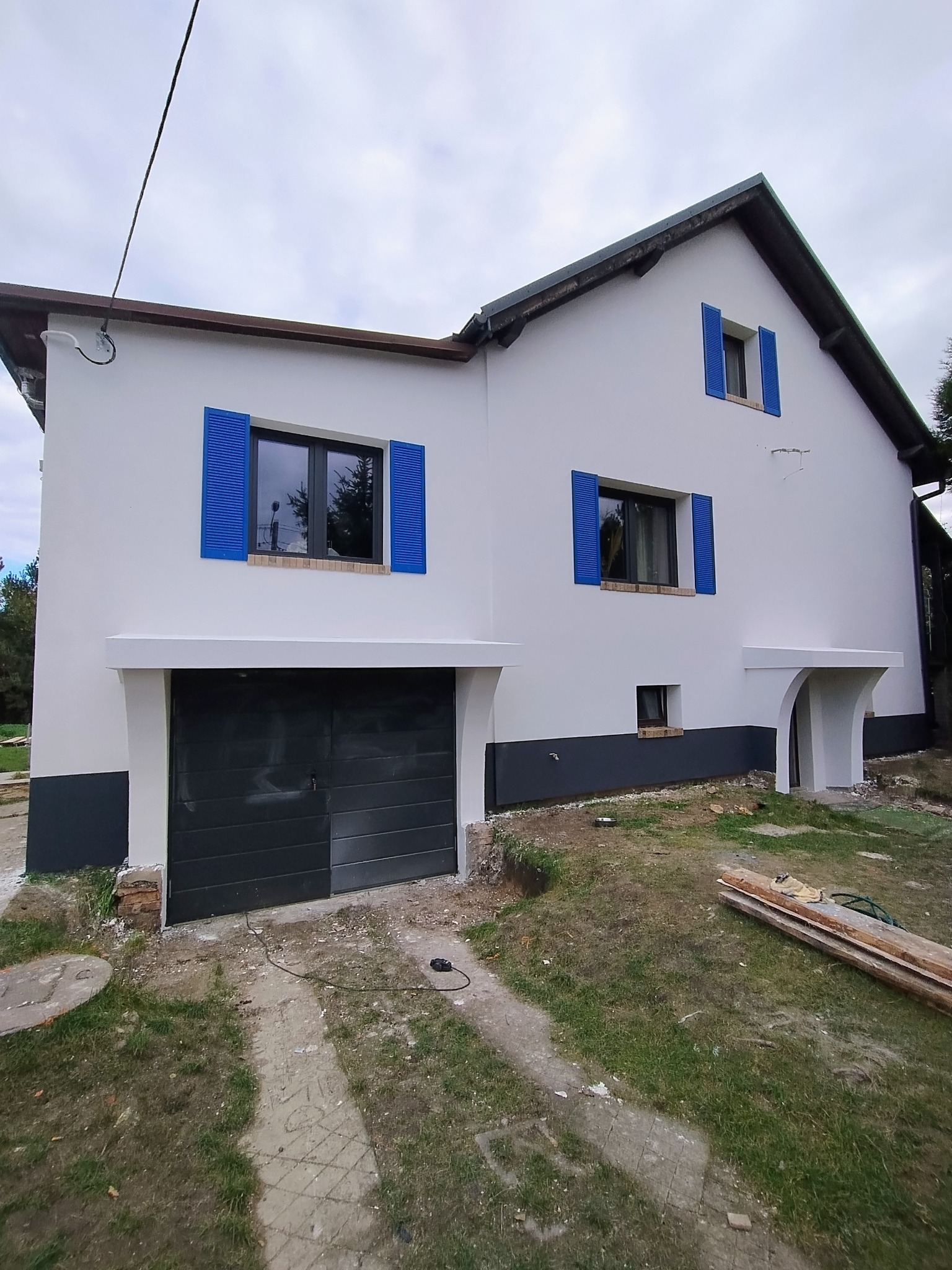 Biały dom po renowacji z niebieskimi okiennicami i ciemnoszarym garażem. Częściowo pomalowane drzwi garażowe. Widok z zewnątrz, w tle pochmurne niebo.