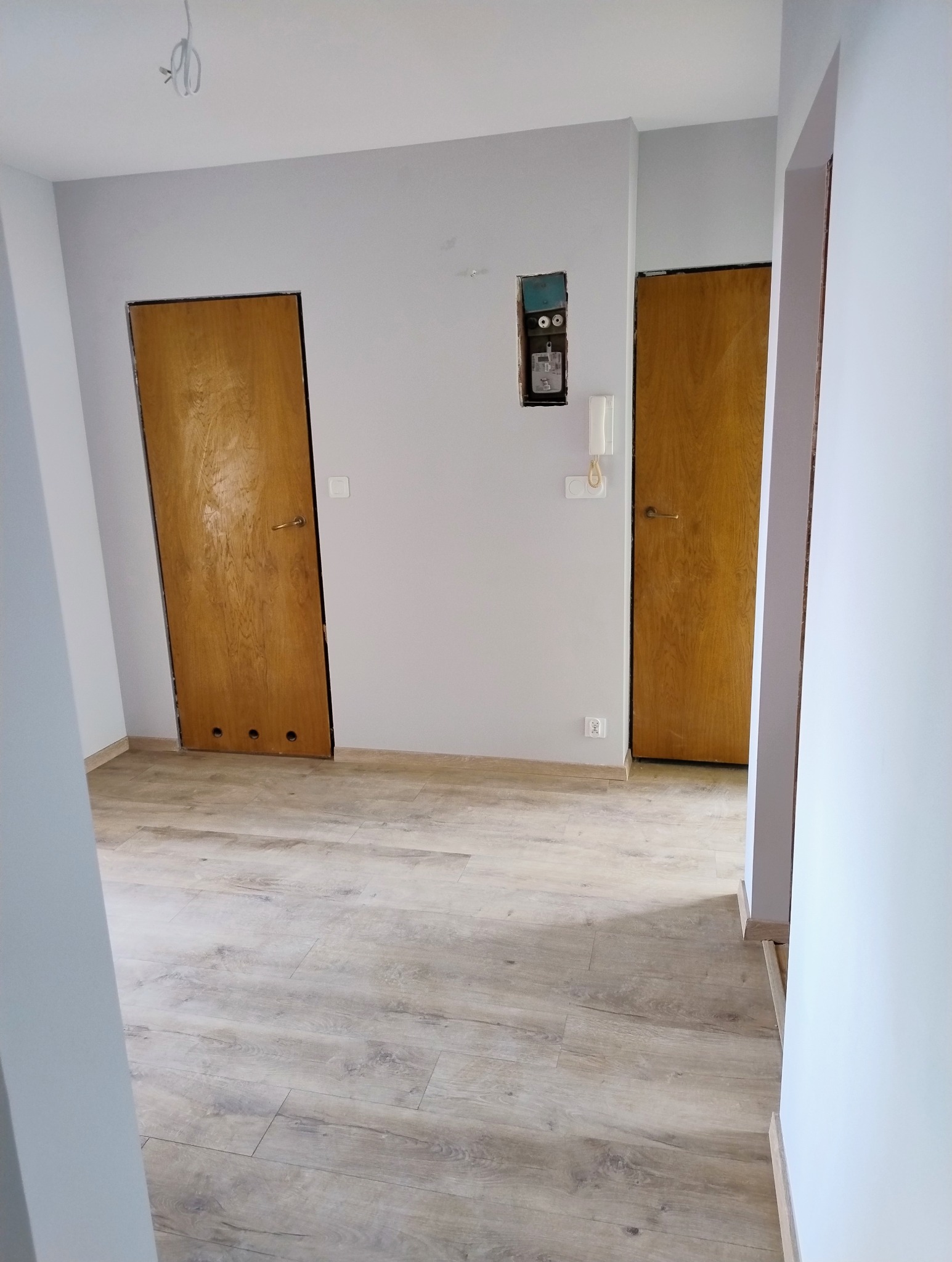 Wnętrze po malowaniu ścian na szaro, z drewnianymi drzwiami i panelami podłogowymi. Widoczna puszka elektryczna bez osprzętu i wystające kable.