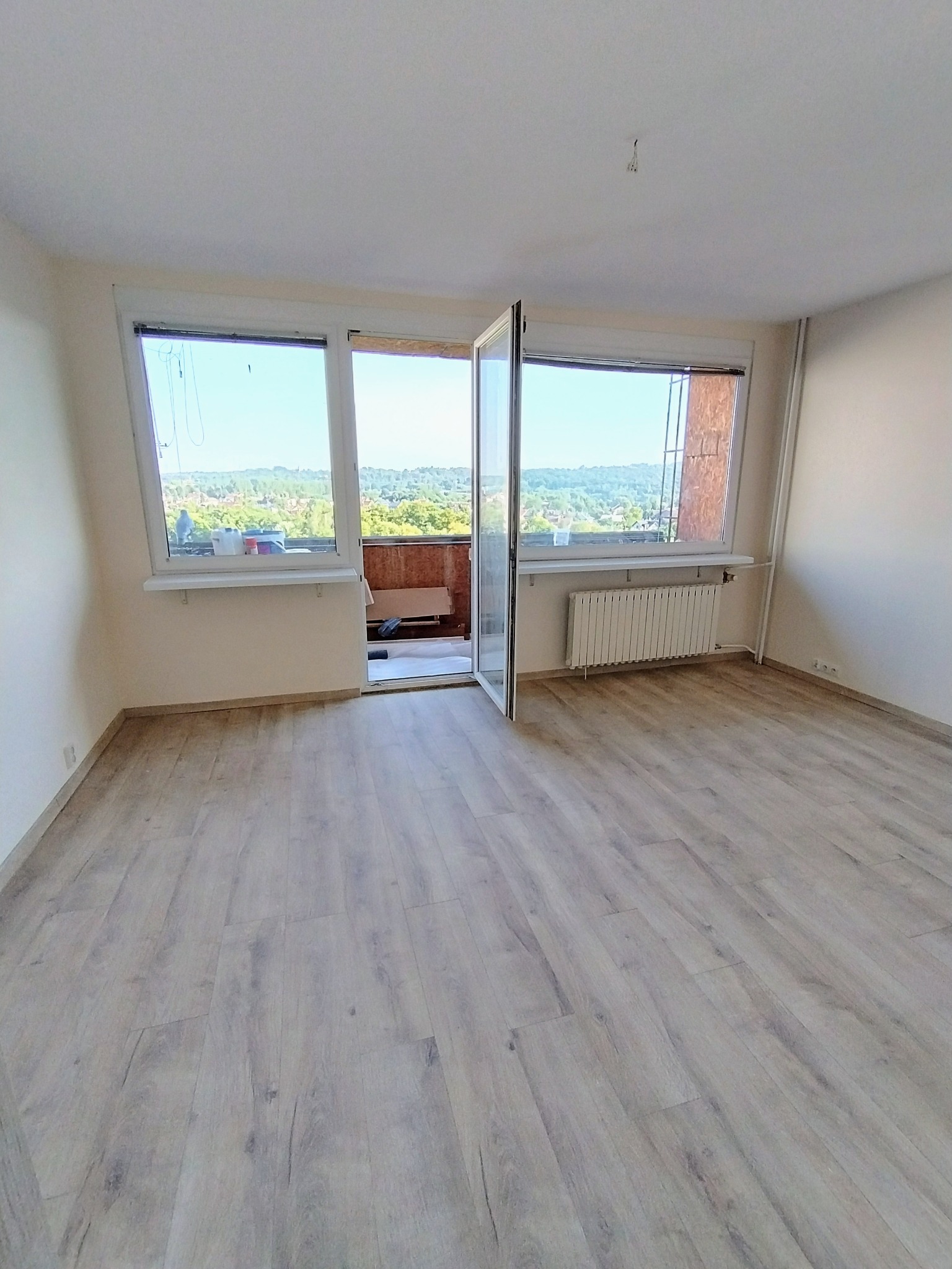 Jasne panele podłogowe w pustym pokoju z otwartym balkonem i widokiem na zieleń. Świeżo wyremontowane wnętrze z grzejnikiem pod oknem.