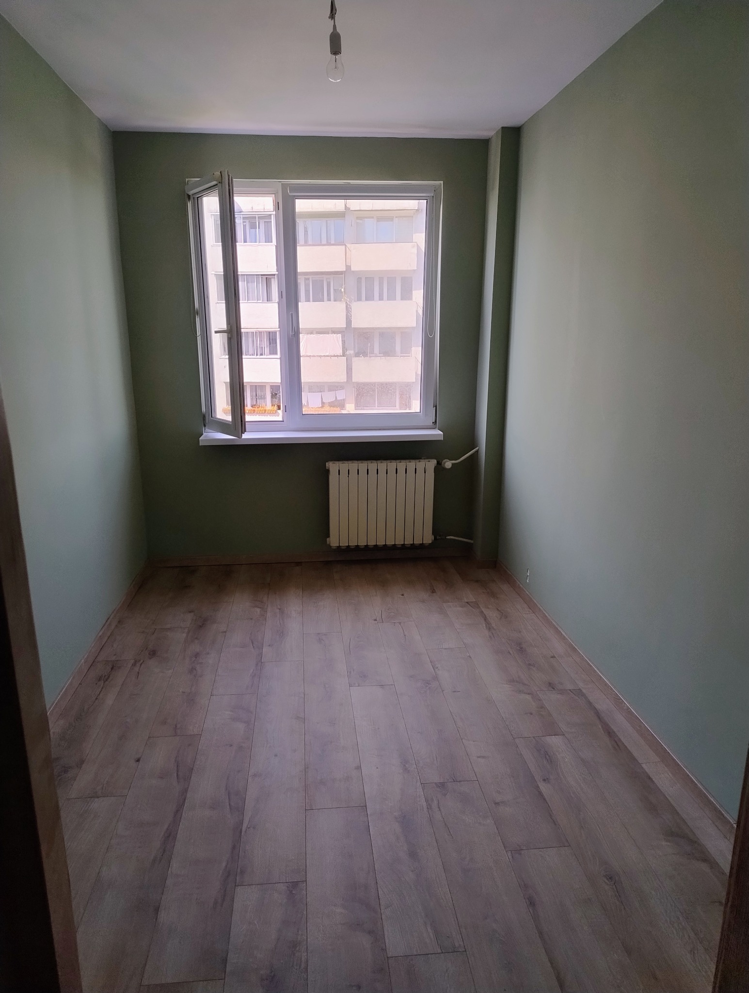 Pusty pokój po malowaniu ścian na oliwkowy kolor, z drewnianą podłogą, białym oknem i grzejnikiem. Widok na budynek z zewnątrz.