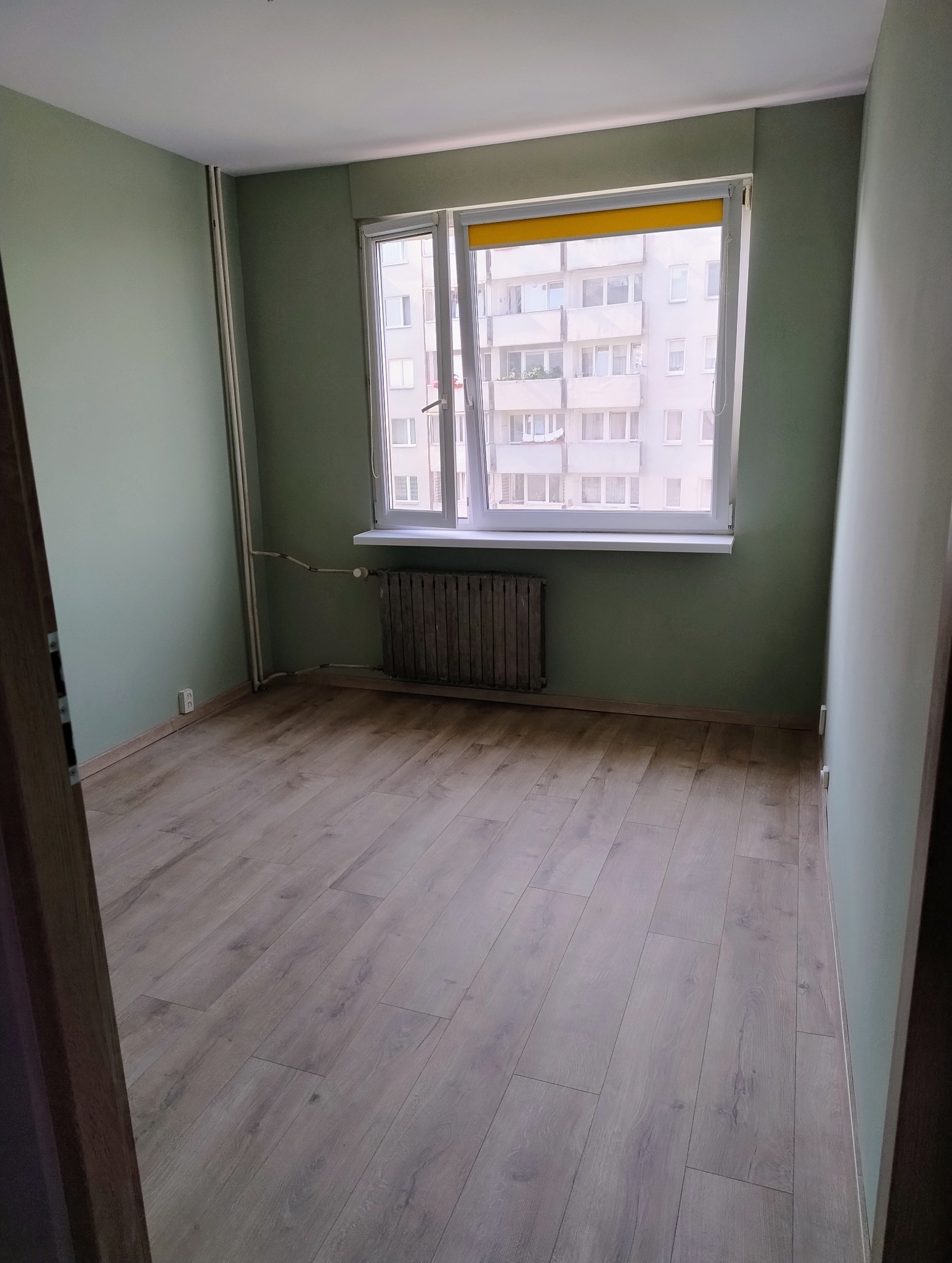Pusty pokój po remoncie: świeżo pomalowane ściany w odcieniach zieleni, panele podłogowe imitujące drewno, grzejnik pod oknem z widokiem na blok.