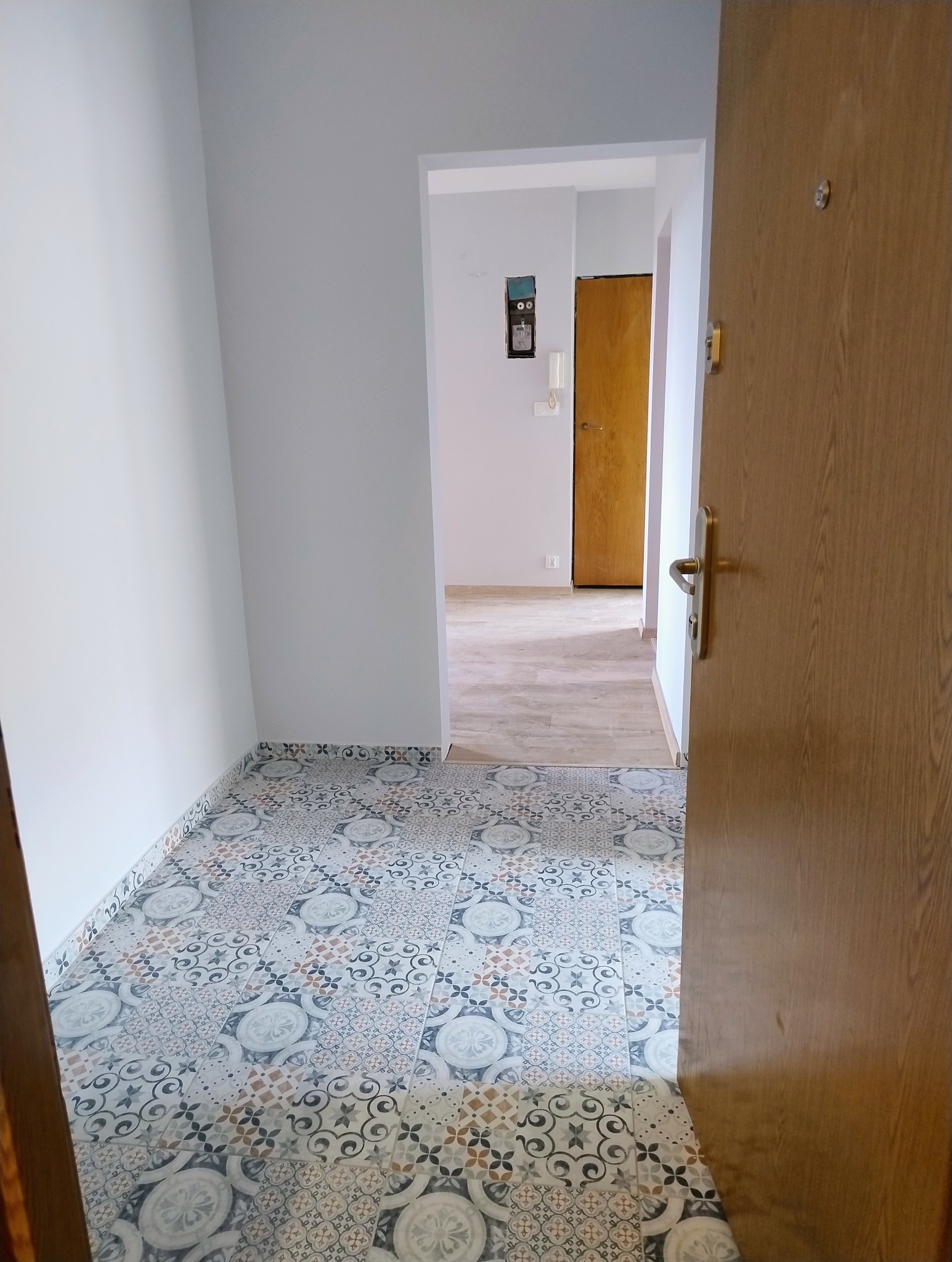 Ukończony remont korytarza: wzorzysta, nowa podłoga z płytek, świeżo pomalowane ściany na szaro, widok na dalszą część mieszkania przez otwarte drzwi.
