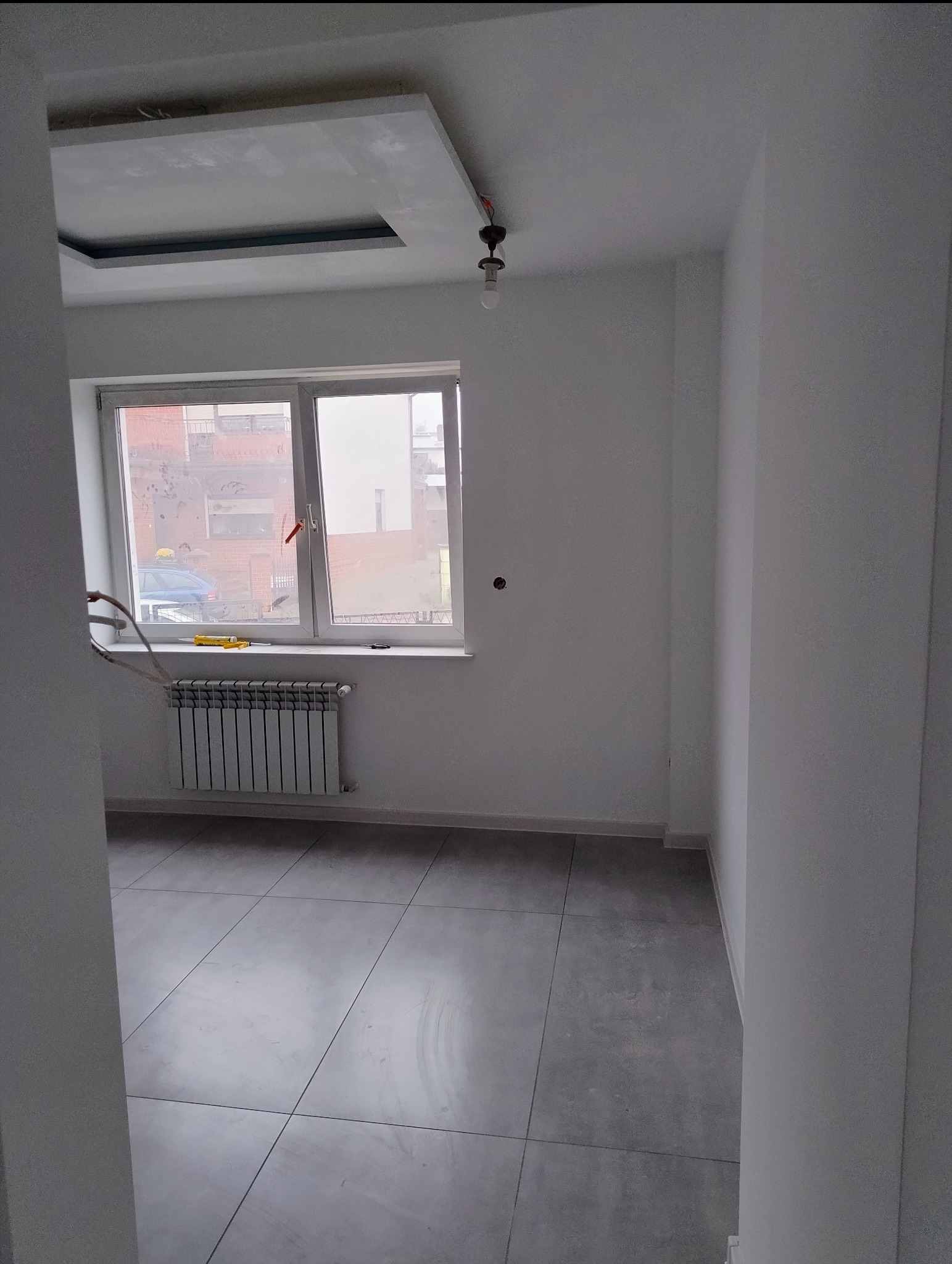 Wnętrze po remoncie: szare płytki podłogowe, białe ściany, okno z grzejnikiem i narzędziami, sufit podwieszany z oświetleniem. Widok zza framugi.