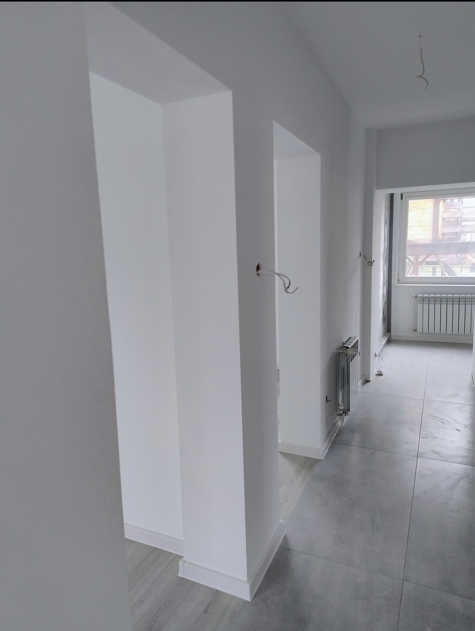 Białe, minimalistyczne wnętrze po remoncie: korytarz z widocznymi elementami instalacji elektrycznej i grzejnikiem. Szare płytki podłogowe, białe ściany i futryny.