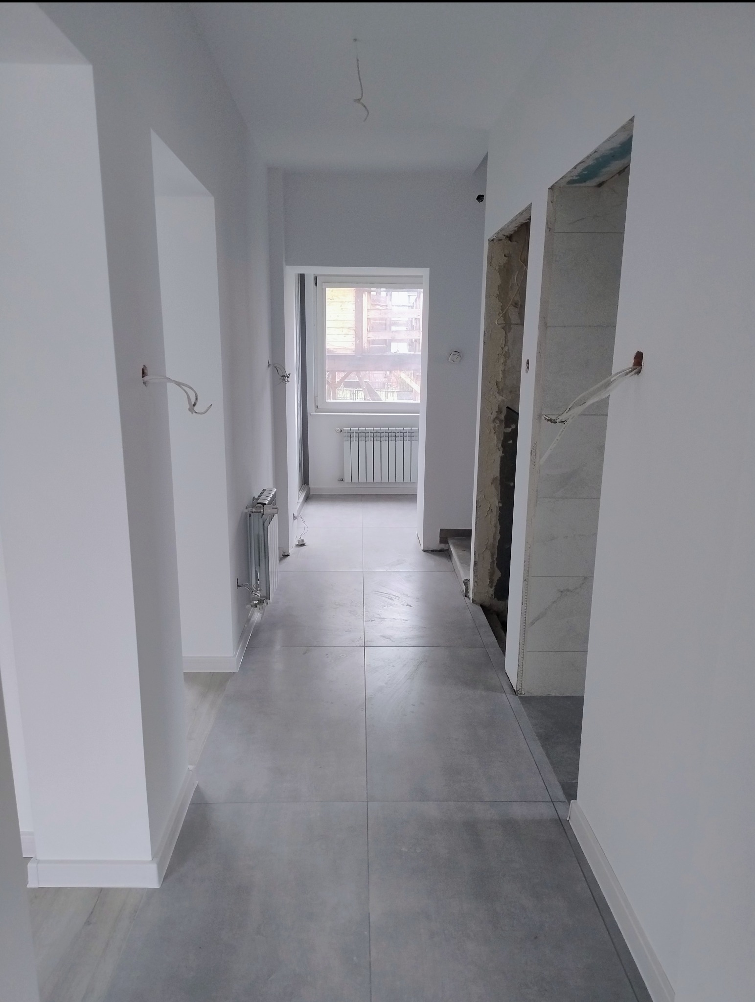 Korytarz w trakcie remontu: szare płytki podłogowe, świeżo pomalowane białe ściany, widoczne instalacje elektryczne i fragmenty ścian przygotowane do wykończenia.