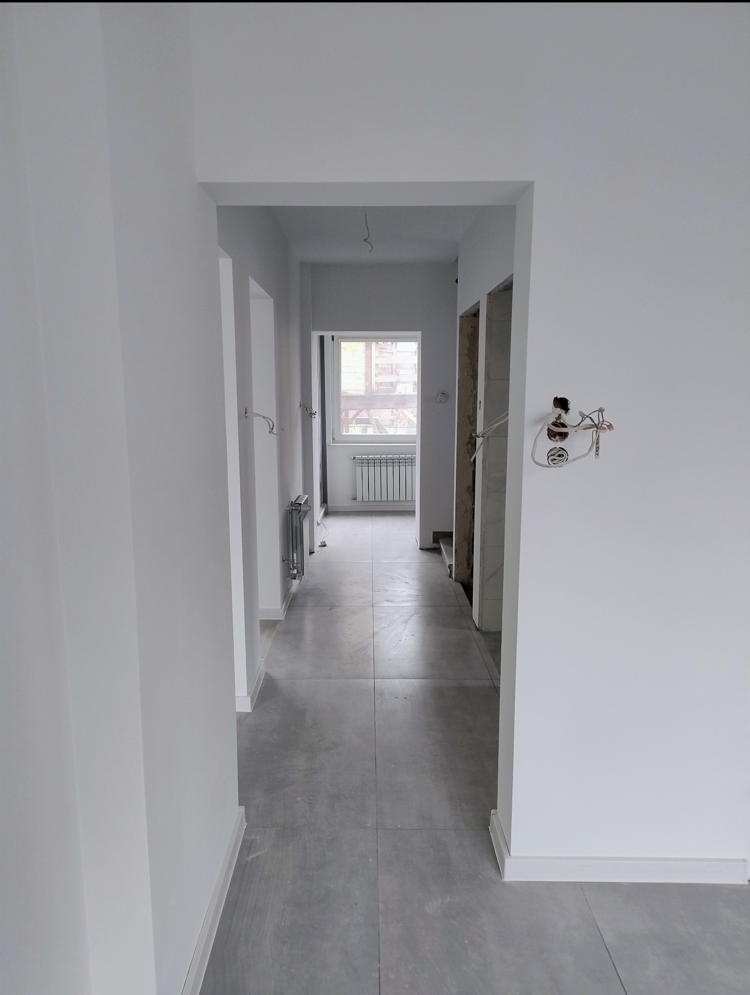 Wnętrze korytarza po remoncie: świeżo pomalowane białe ściany, szara podłoga z płytek, widoczne grzejniki i instalacja elektryczna w trakcie prac.